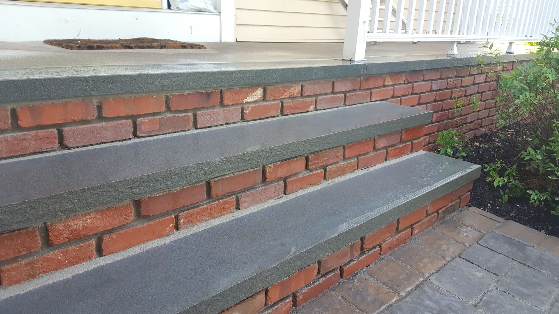 Brick steps with gray concrete treads leading to a porch beside white siding and shrubs