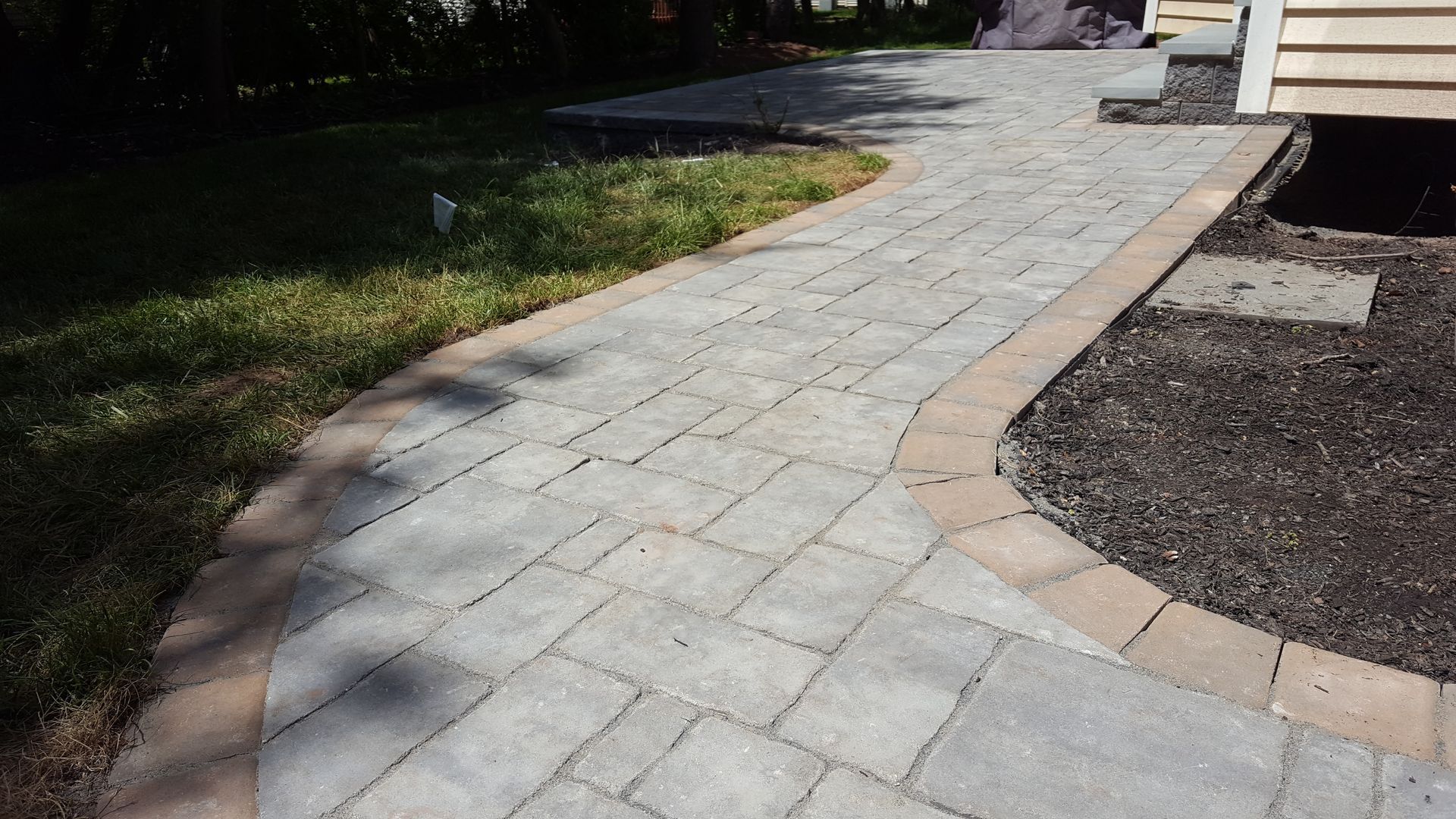 Stone walkway curves past grass and gravel beside a driveway in sunlight
