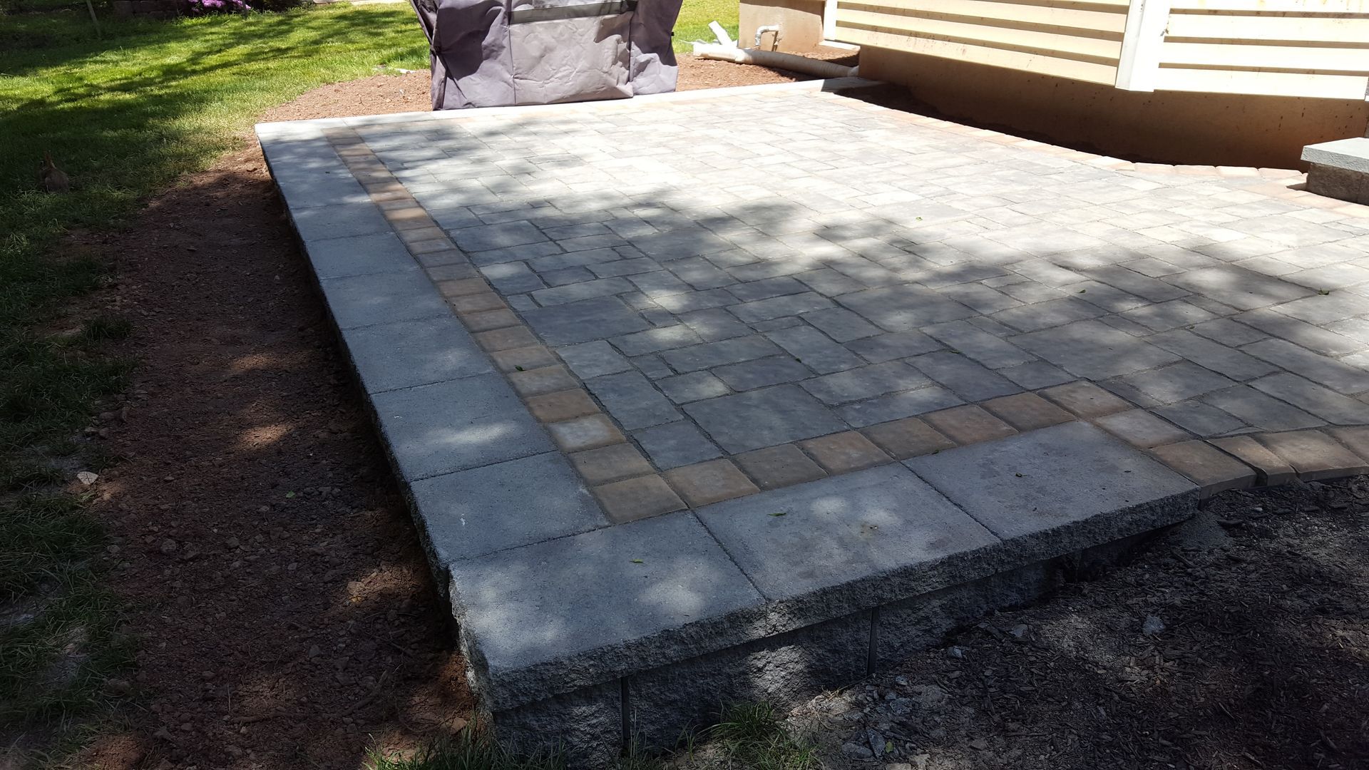 Stone patio with square pavers beside a house, bordered by grass and a small retaining wall