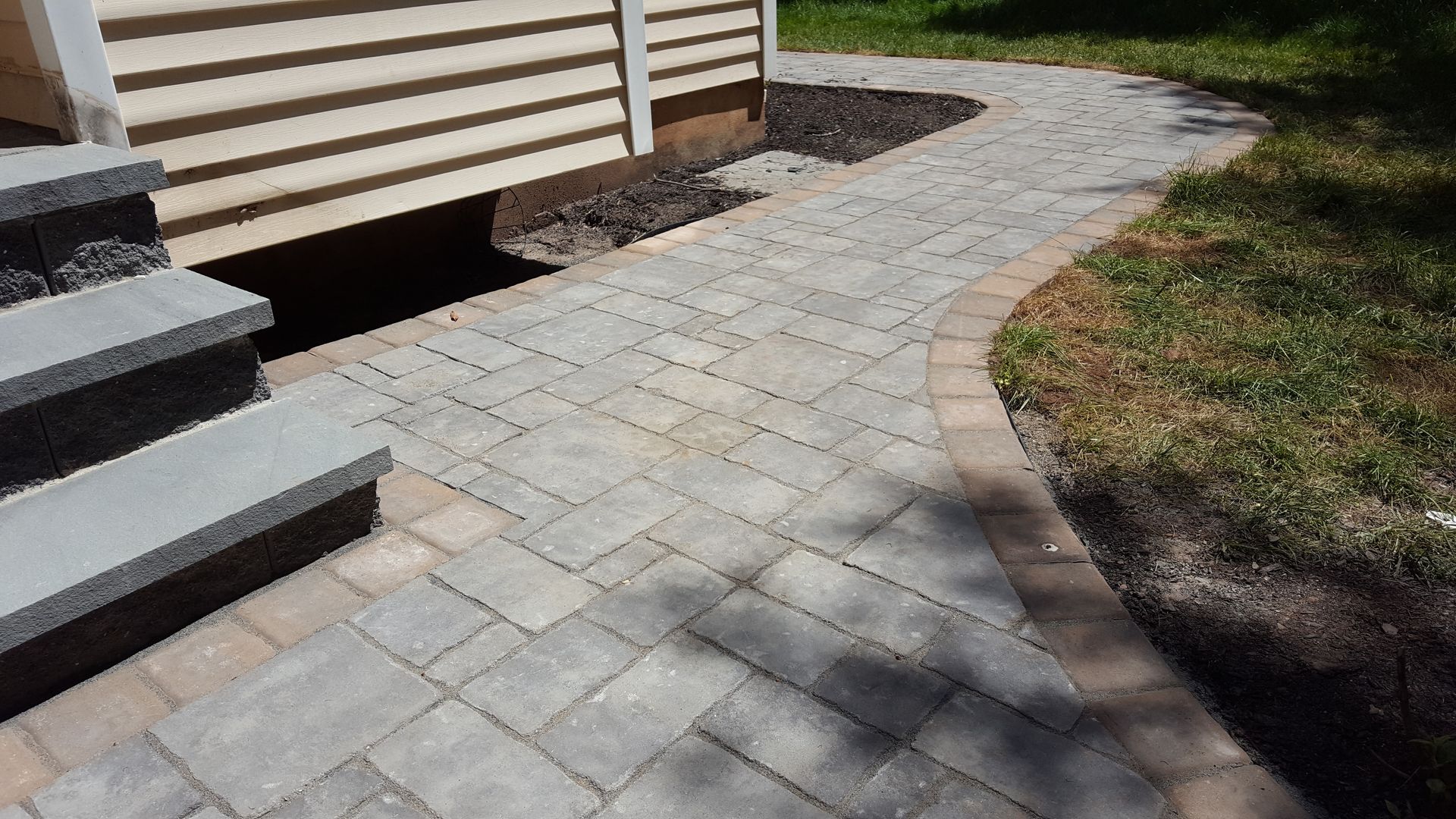 Curved stone paver walkway beside a house and steps, bordered by grass and mulch