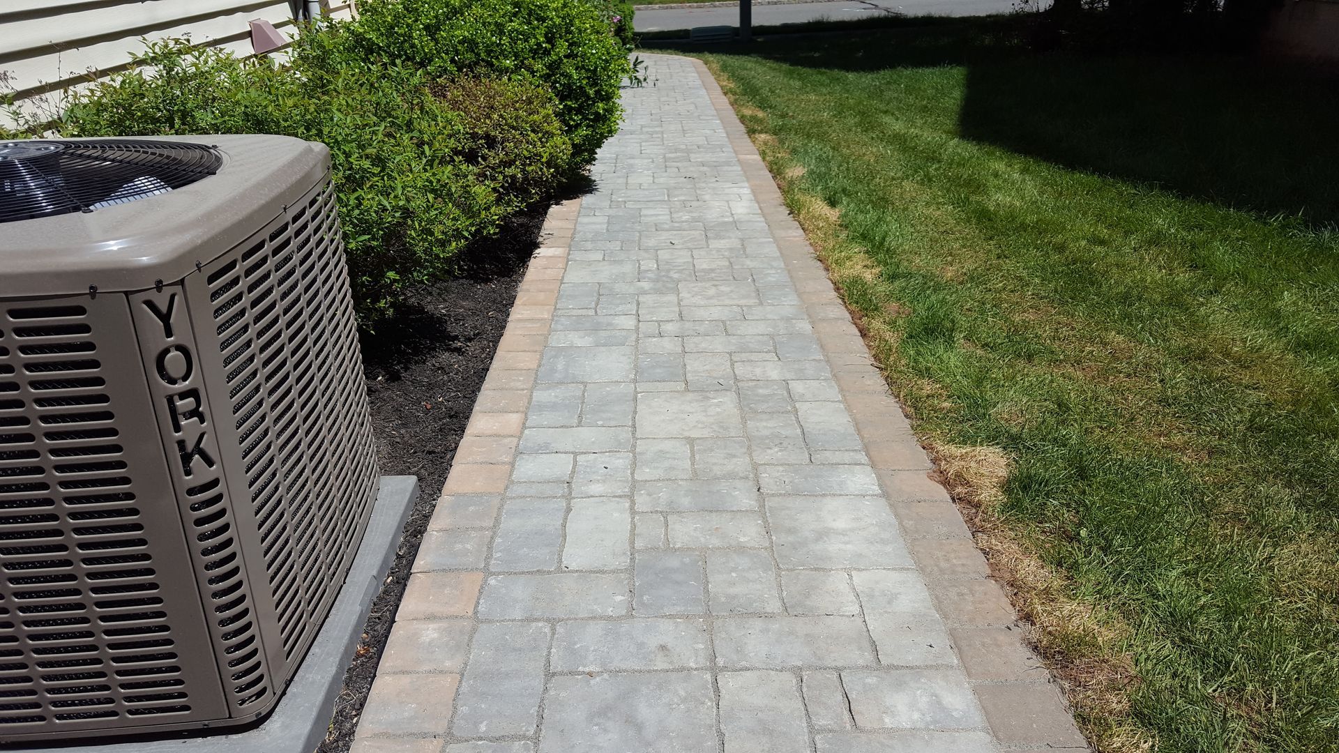 Stone sidewalk beside grass and shrubs next to an outdoor air conditioning unit