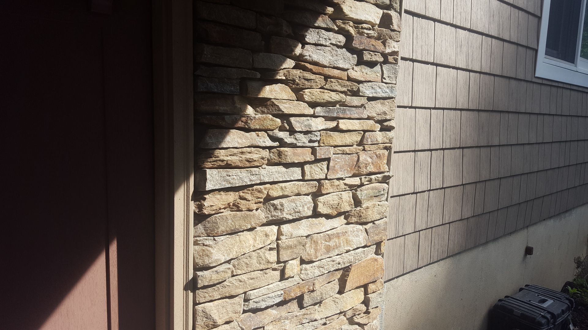 Stone-faced exterior wall beside a dark doorway, with tan shingle siding and sunlight casting shadows.