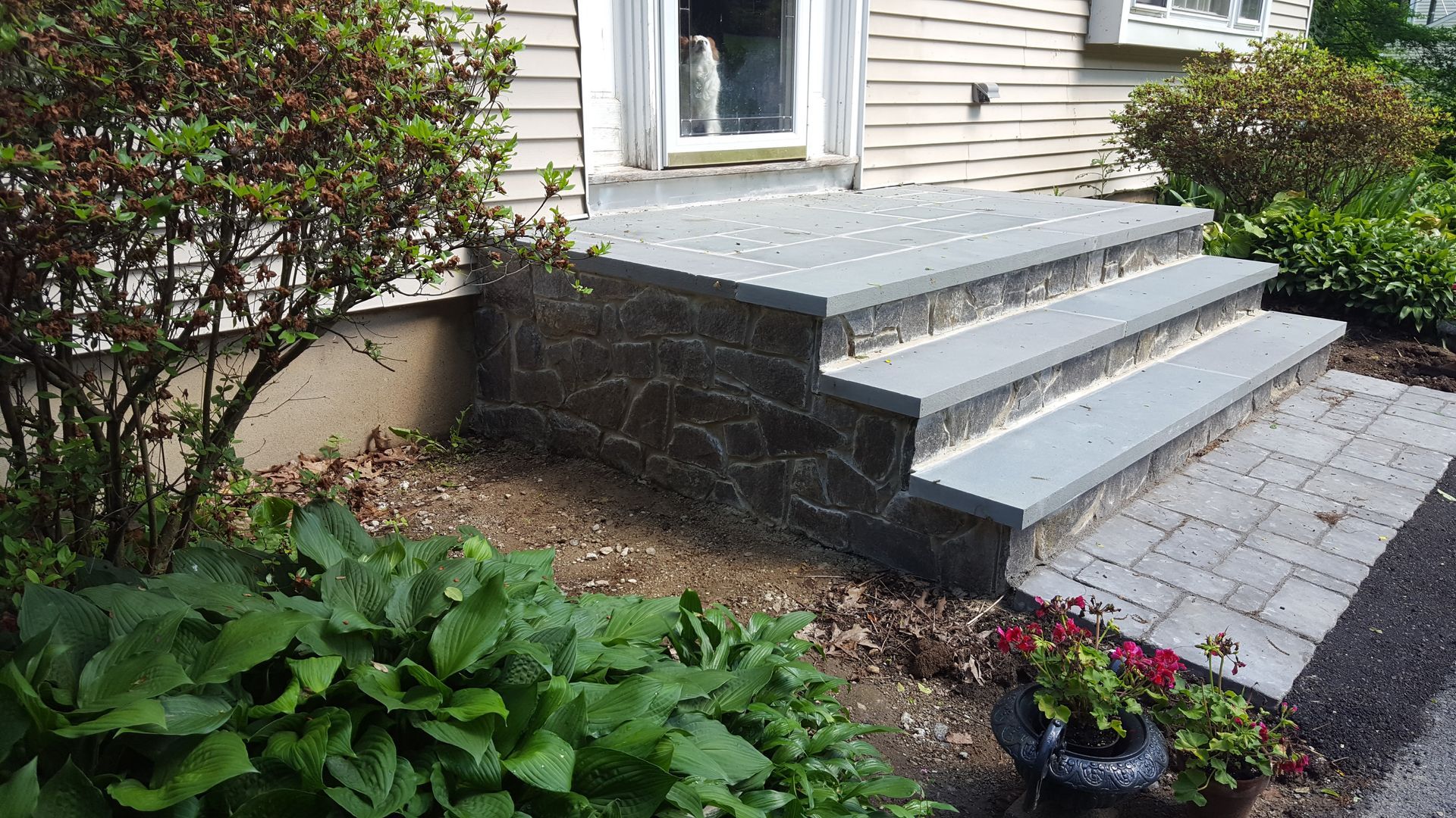 Front steps with gray stone retaining wall, light patio pavers, and flower beds by a house entrance