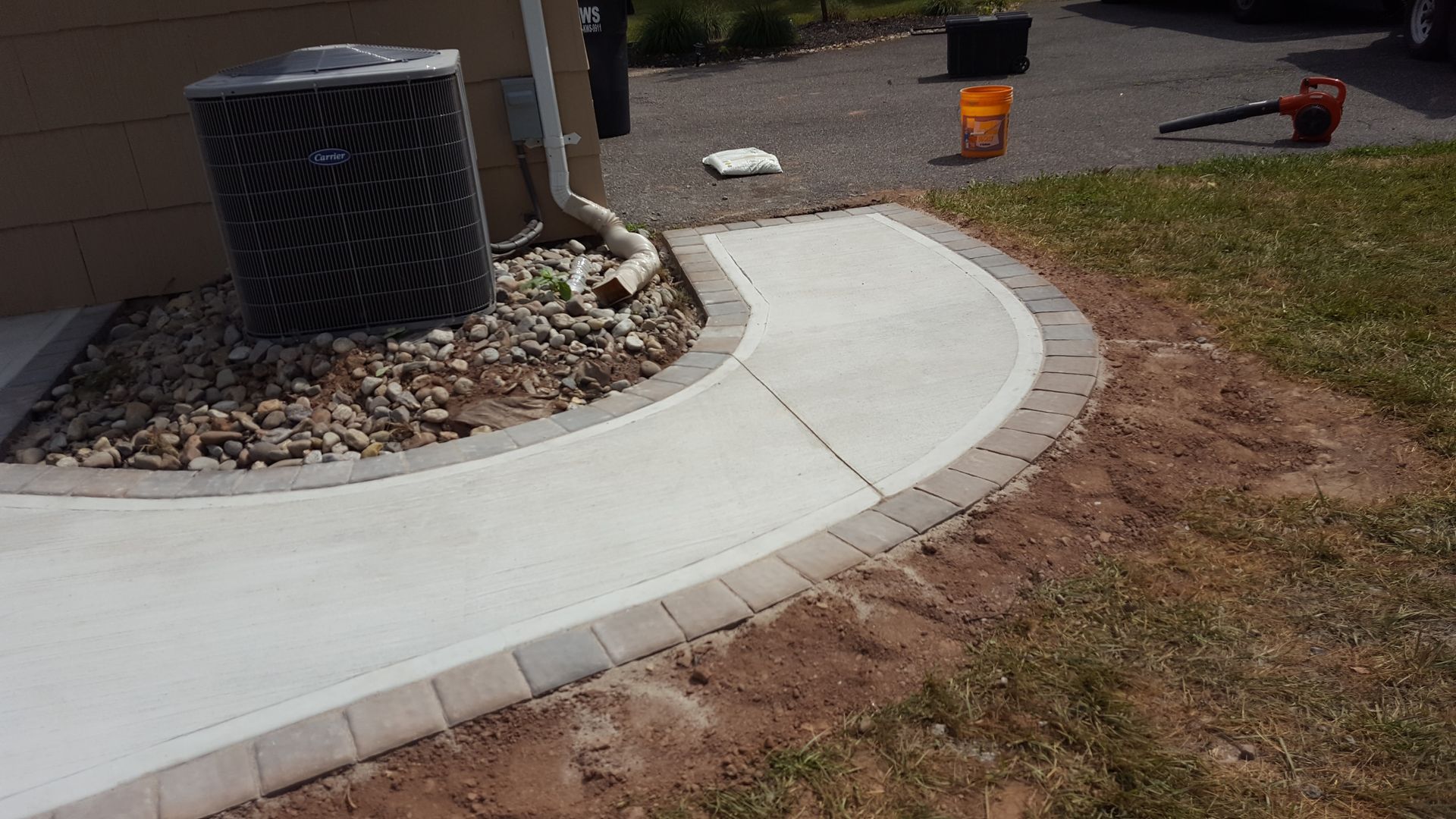 Curved concrete walkway beside a house, with an HVAC unit and a wheelbarrow nearby.