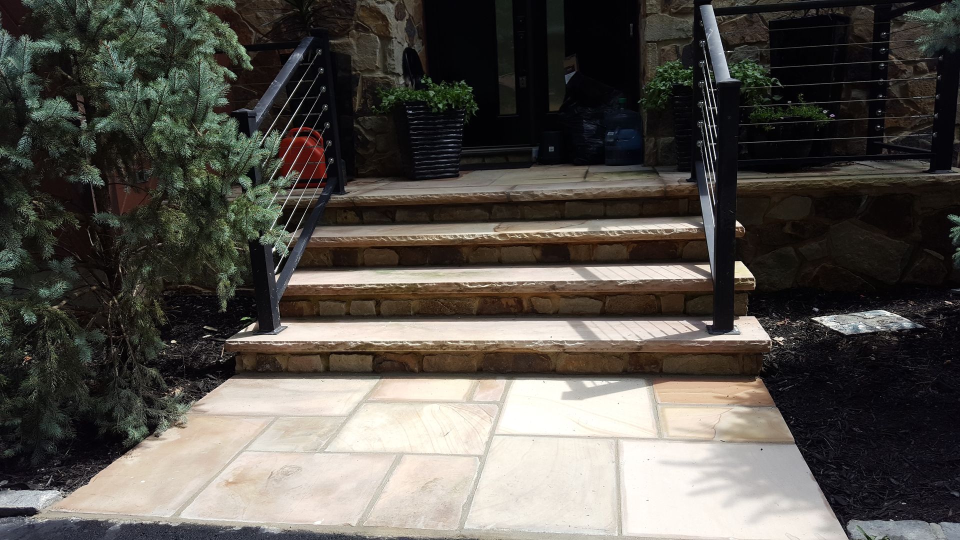 Stone steps leading to a shaded patio entrance with a black handrail and potted greenery