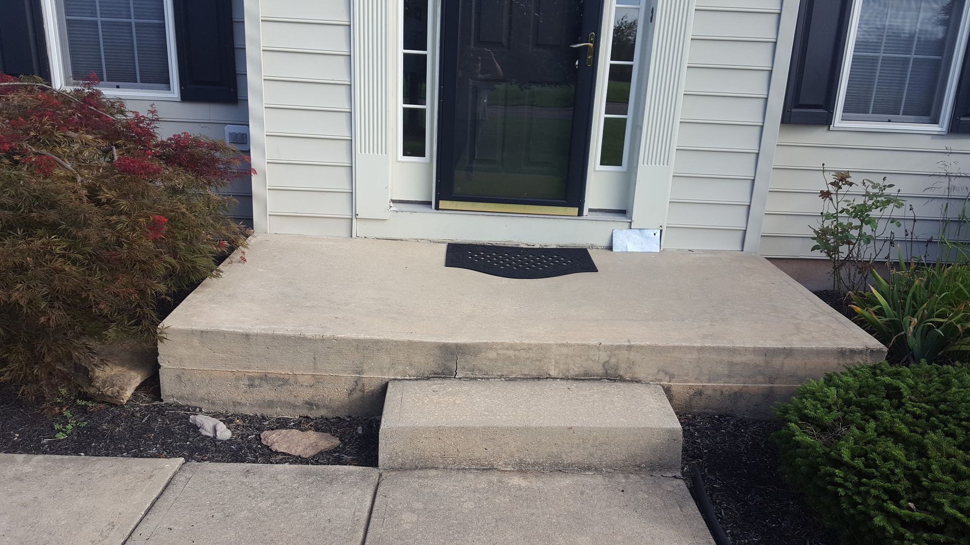 Front porch with concrete steps and landing leading to a black front door, flanked by white siding and shrubs.