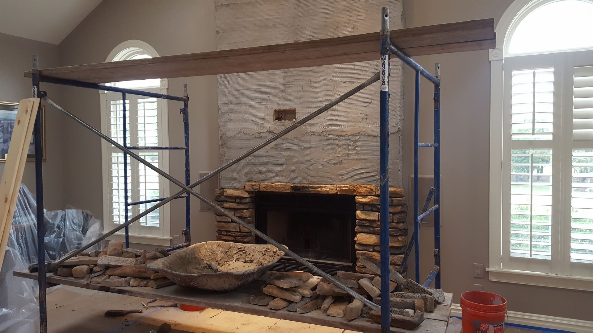 Scaffolded fireplace wall under renovation with stonework, wooden mantel, and window shutters to the right