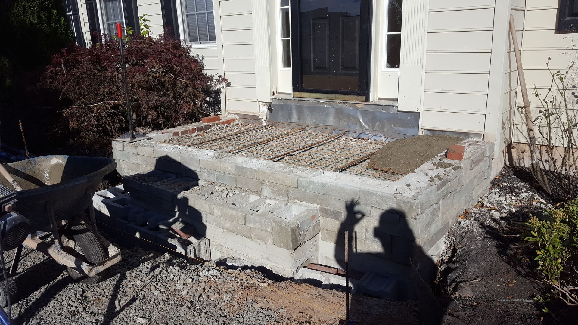Front porch under construction with a newly poured concrete step and a wheelbarrow beside the house.