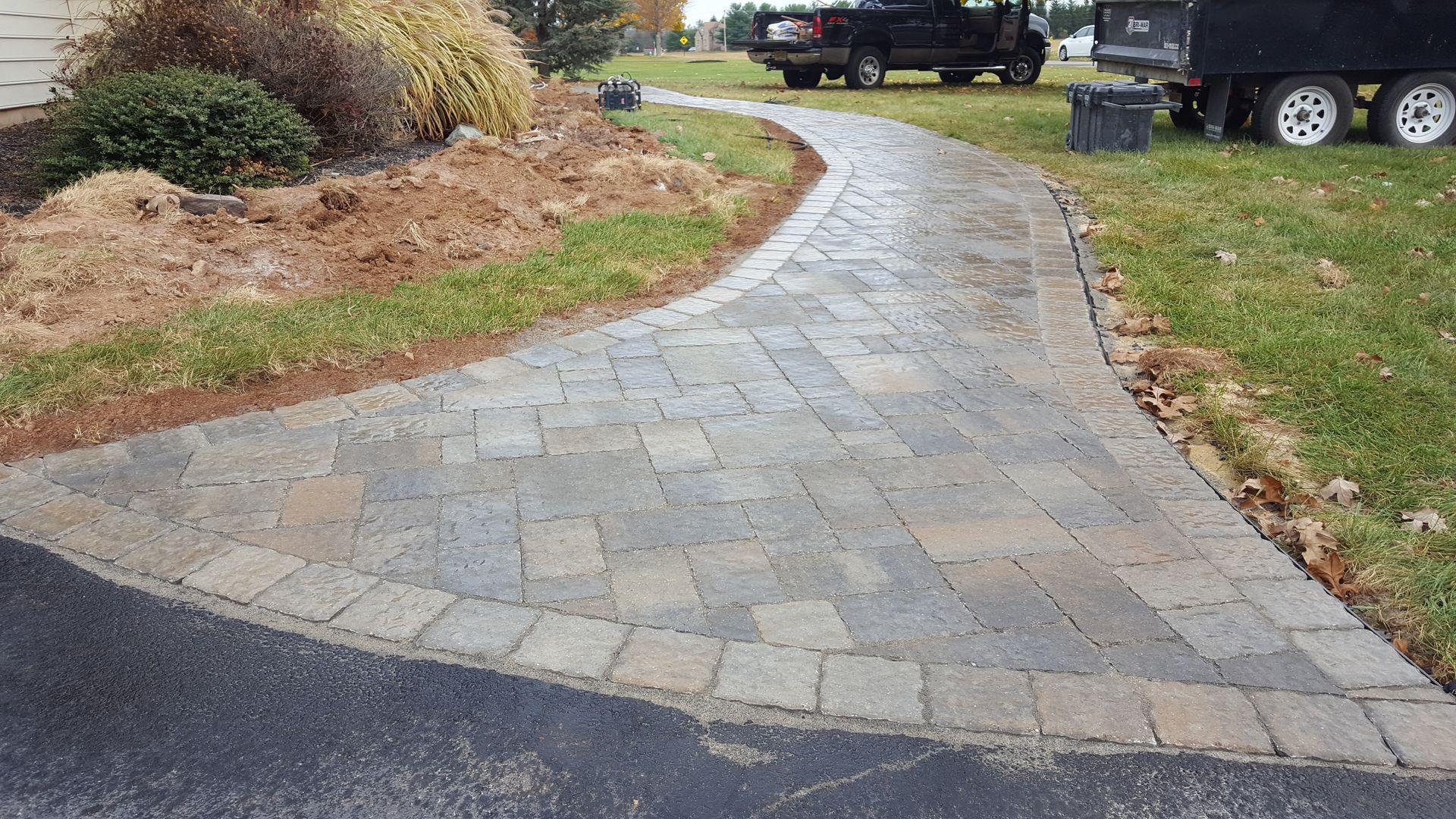 Curved stone paver walkway through a landscaped yard beside a driveway and parked truck
