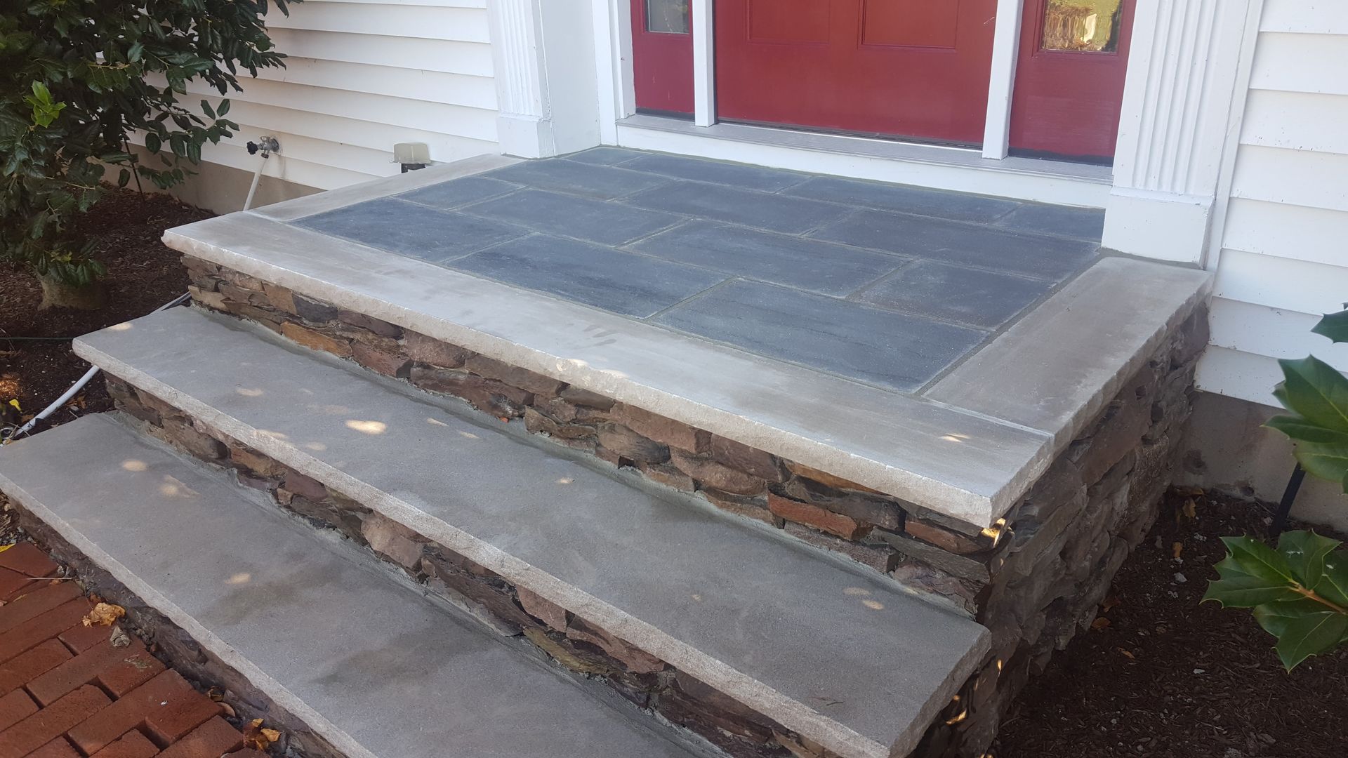 Front steps and landing leading to a red door, with cracked concrete and exposed brick edges.