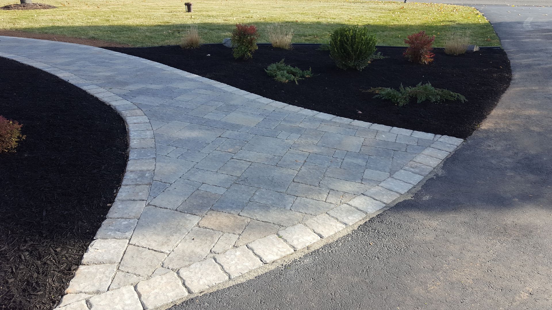 Curved light stone walkway bordered by dark mulch and asphalt driveway.