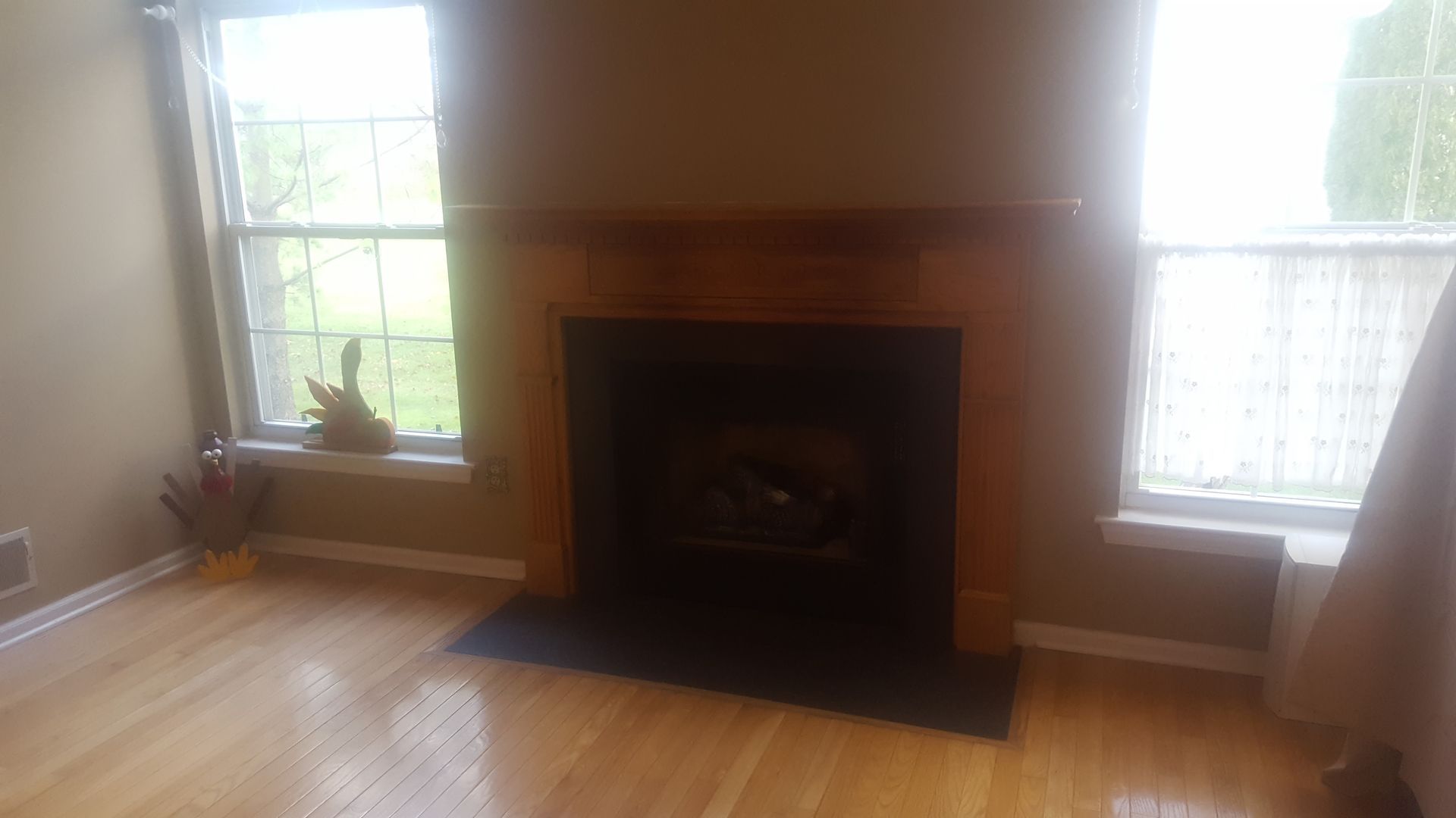 Living room fireplace between two bright windows, with hardwood floors and a wooden mantel.