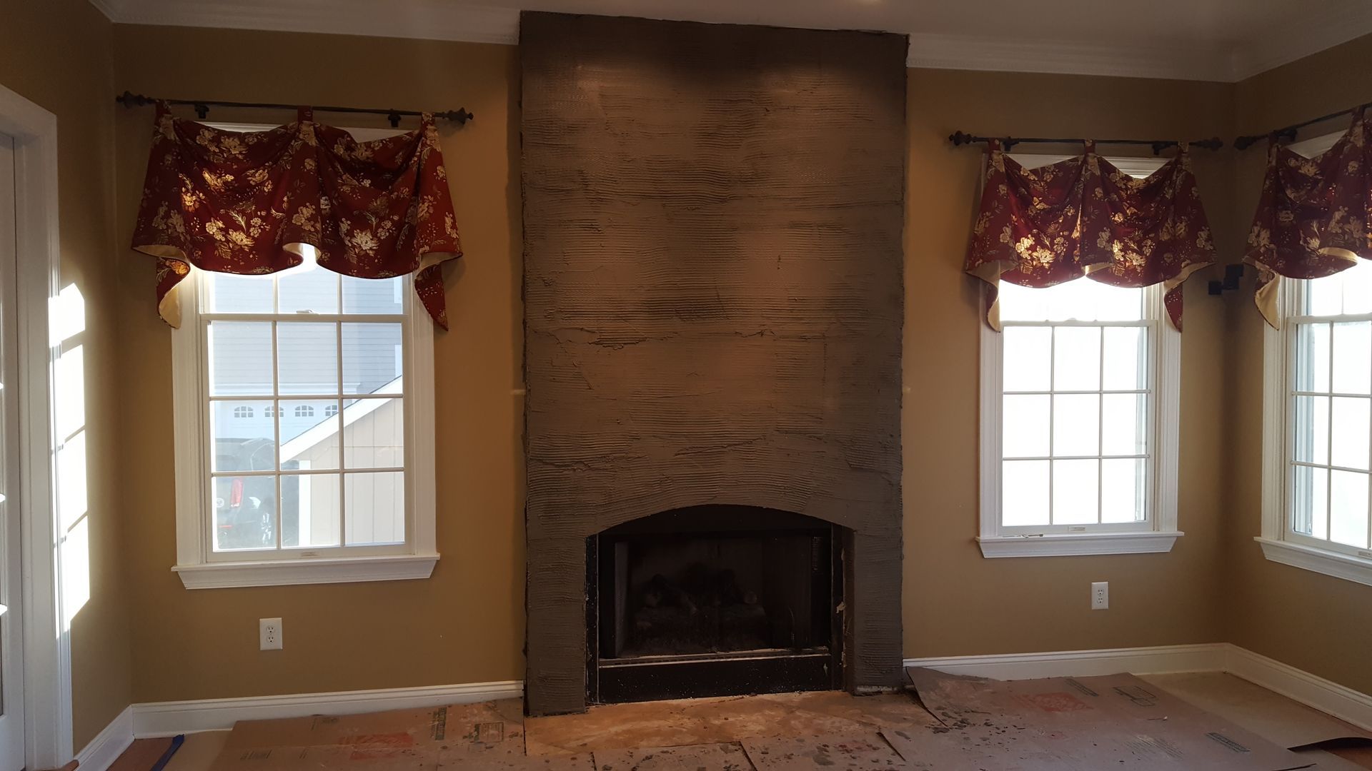 Empty living room with a central brick fireplace, two windows, and brown walls with floral valances