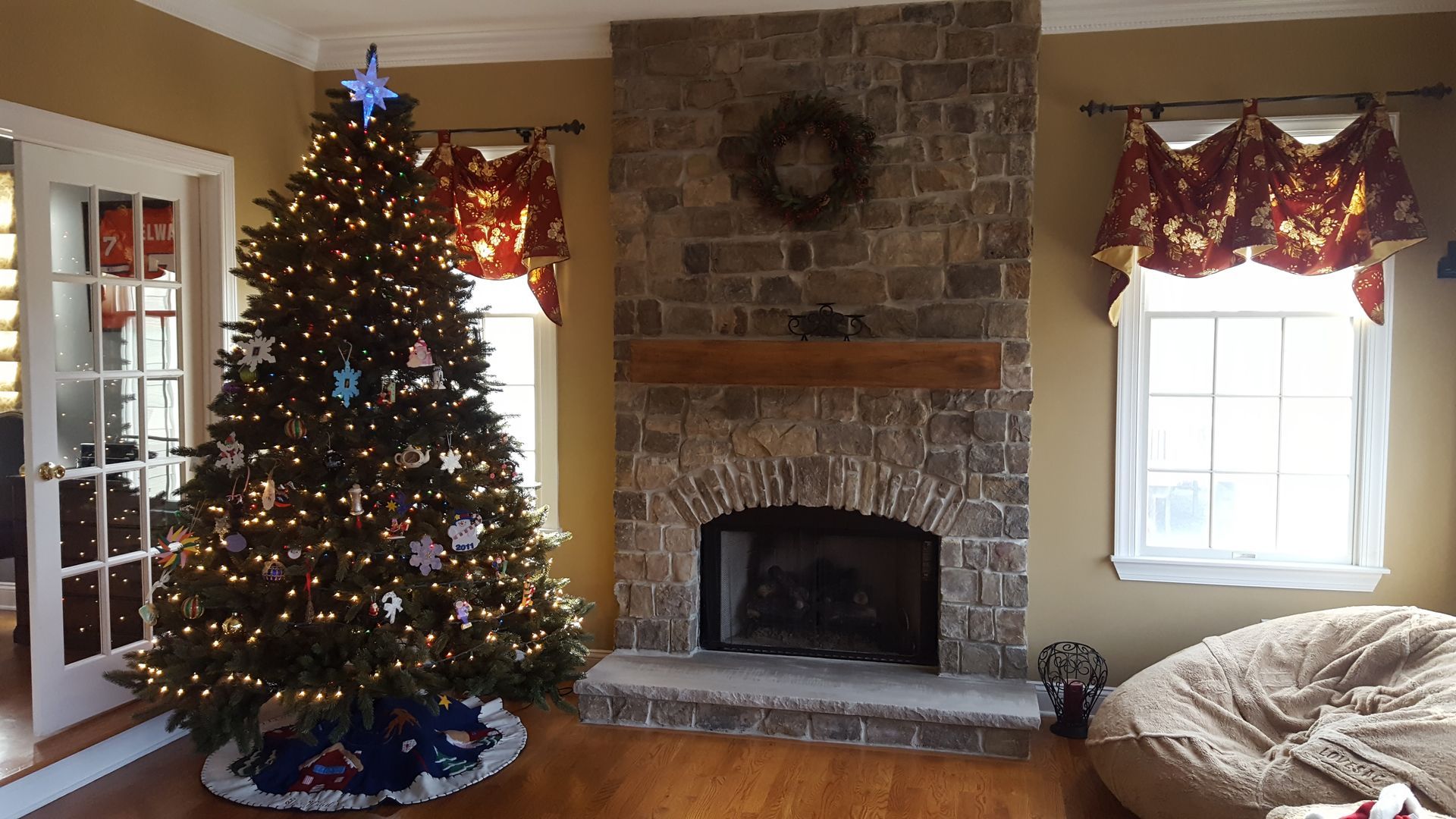 Christmas tree in a cozy living room beside a stone fireplace and decorated windows