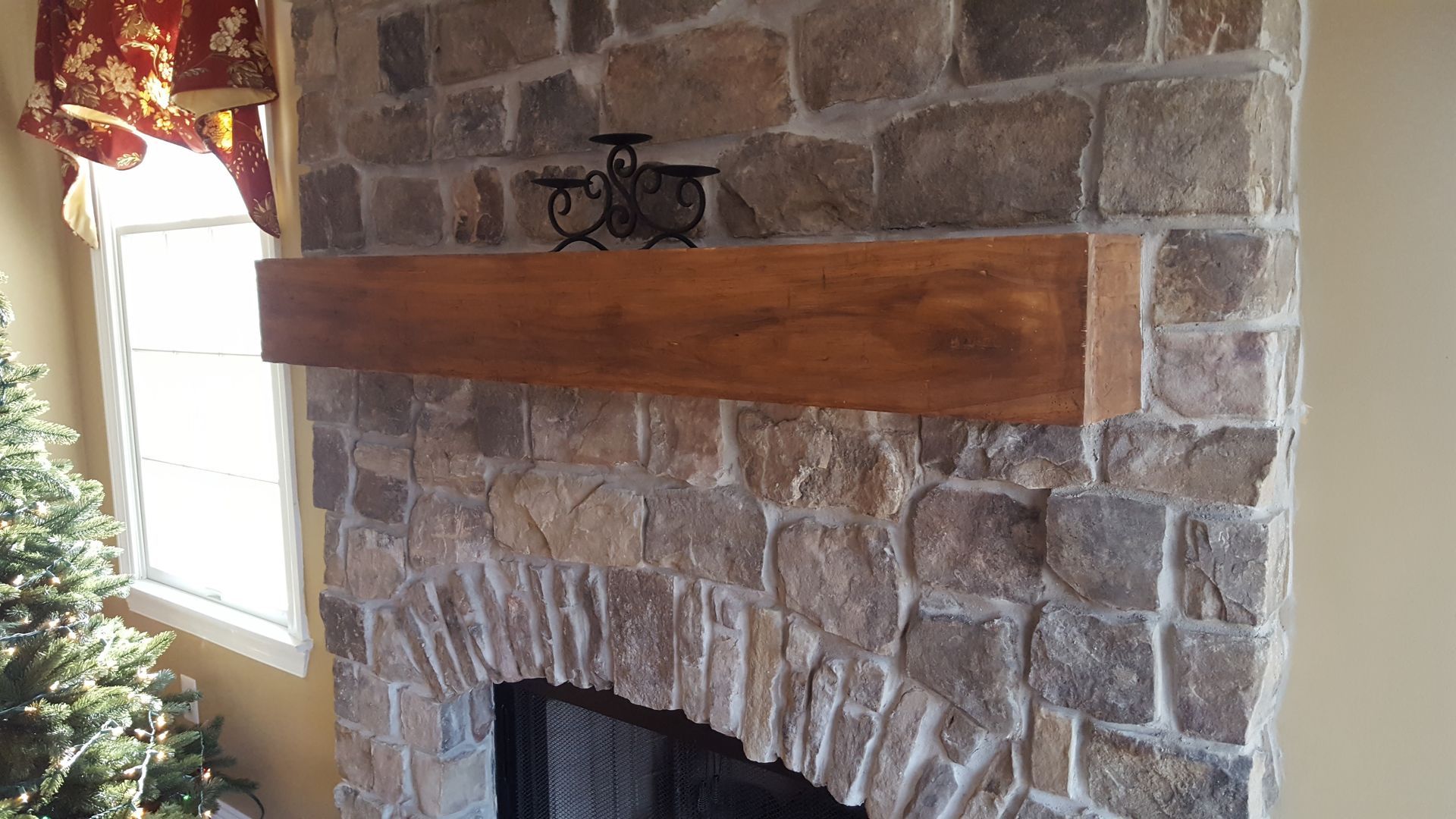 Stone fireplace with a thick wooden mantel in a living room, beside a bright window and red curtain