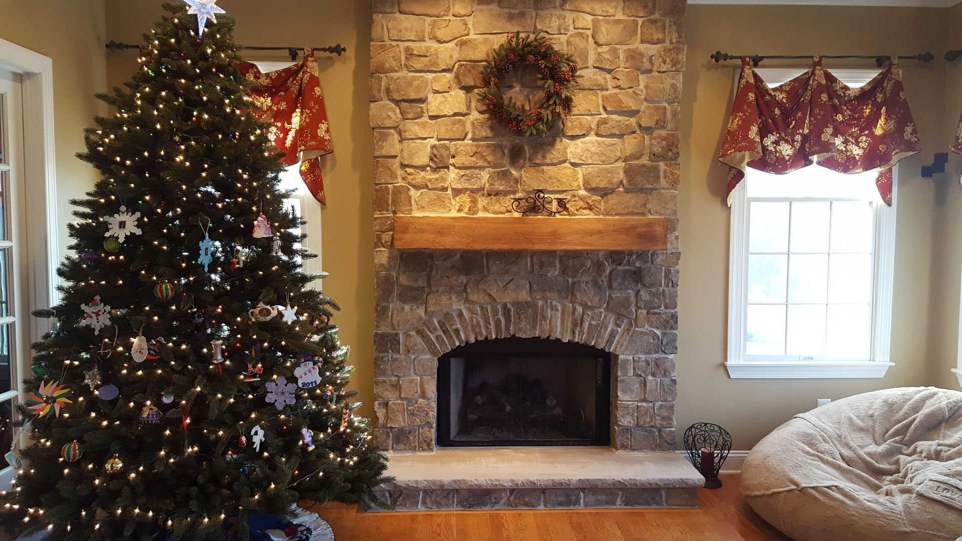 Christmas tree beside stone fireplace in cozy living room with wreath, window valance, and beanbag chair