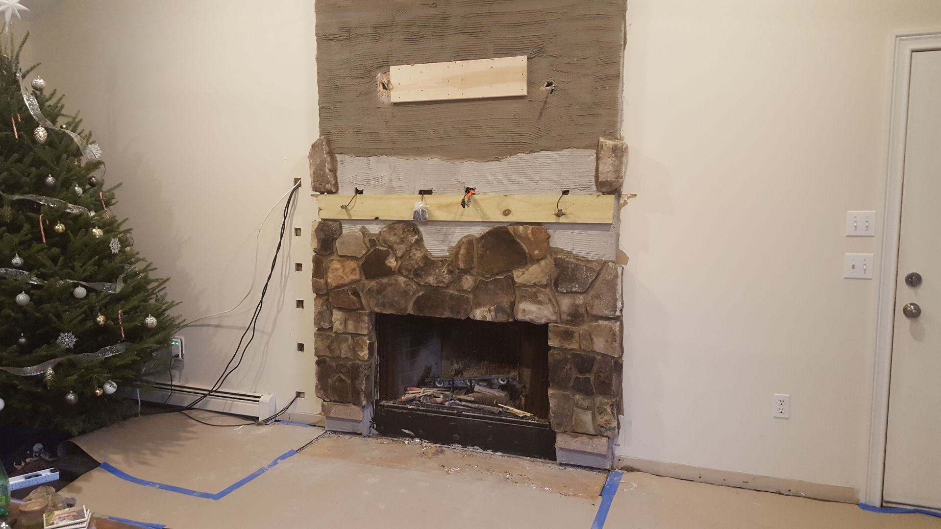 Living room with a stone fireplace under renovation, blue tape on the carpet, and a Christmas tree at left