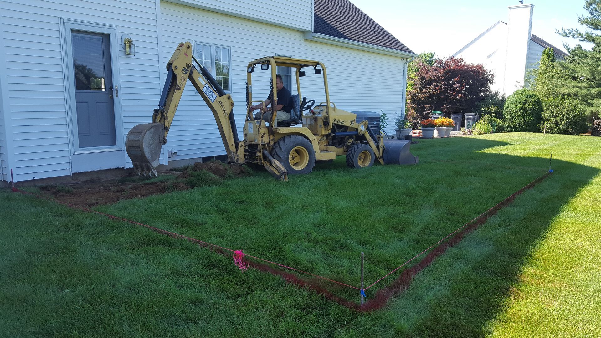 Backhoe excavator beside a house, digging a red-outlined section of lawn