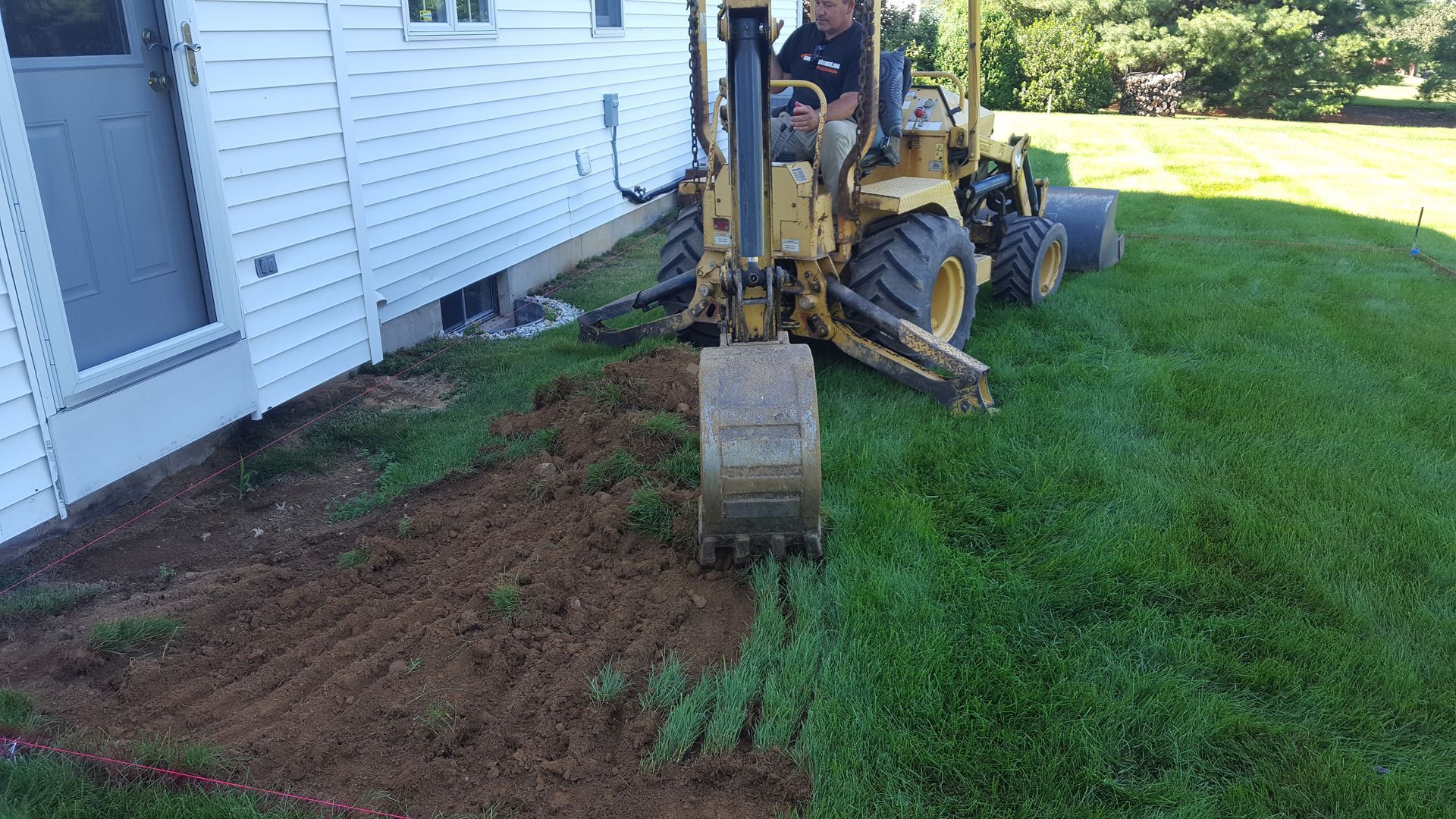 Backhoe digging a trench beside a white house, with a worker on the machine and fresh dirt exposed.