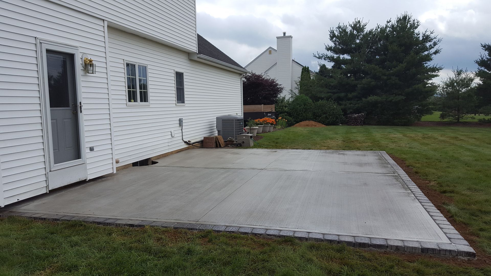 Concrete patio slab beside a white house, with a grassy yard and trees beyond.
