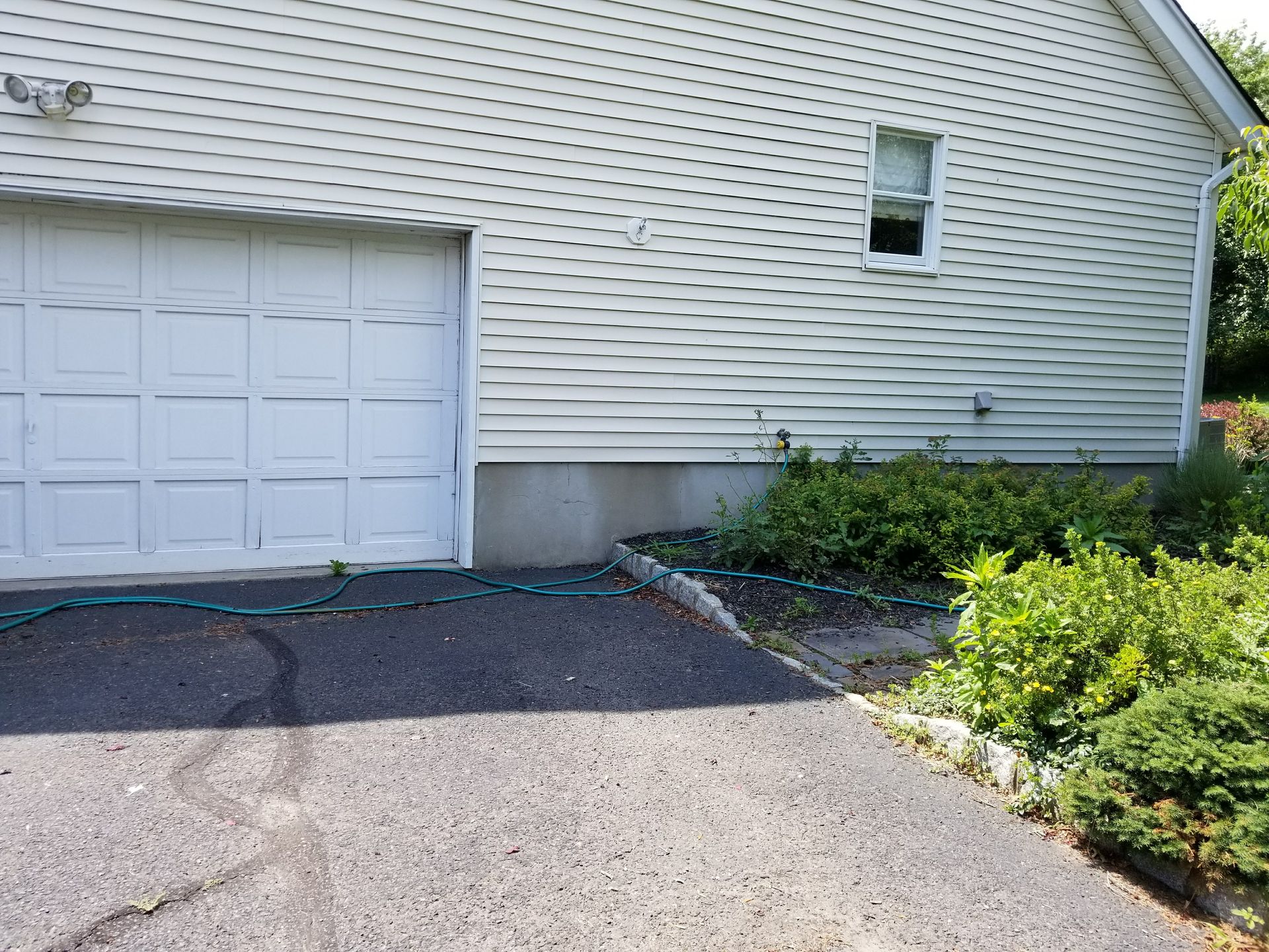 White garage with driveway, siding, and shrubs beside a house exterior