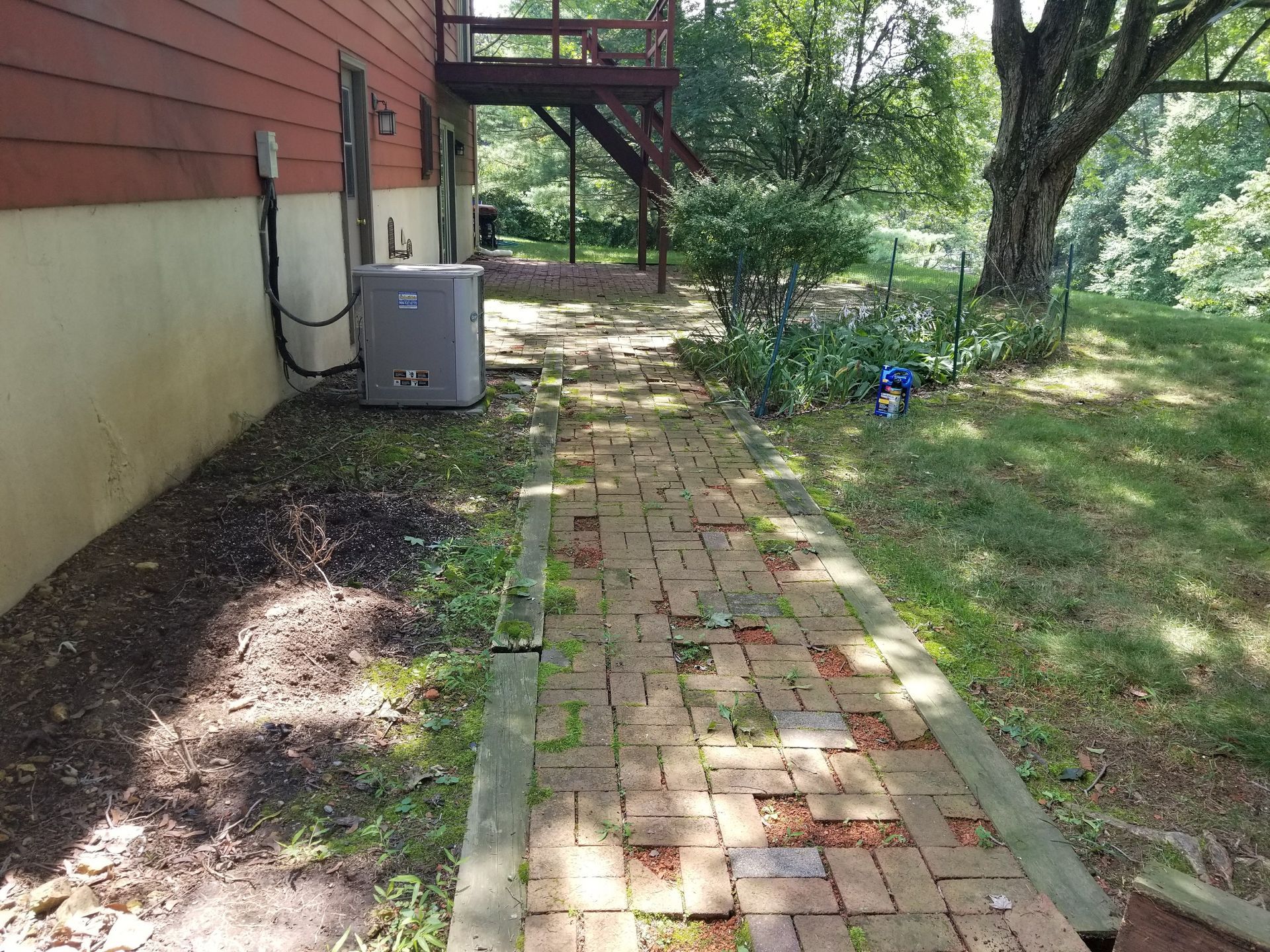 Brick walkway beside a house with an HVAC unit, trees, and a small blue garden item in a yard