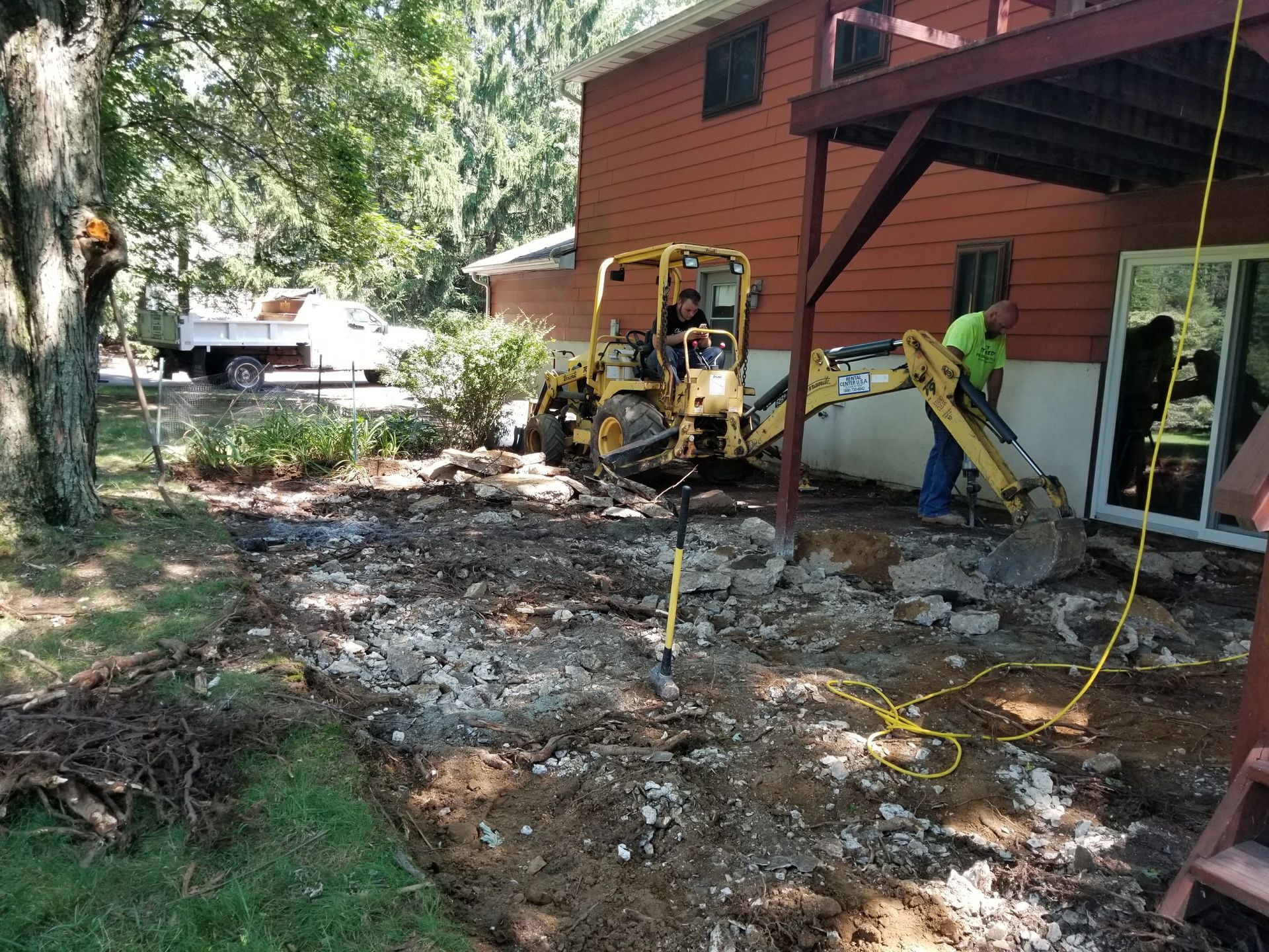 Backyard construction scene with a yellow excavator, dirt piles, and a red house deck on the right