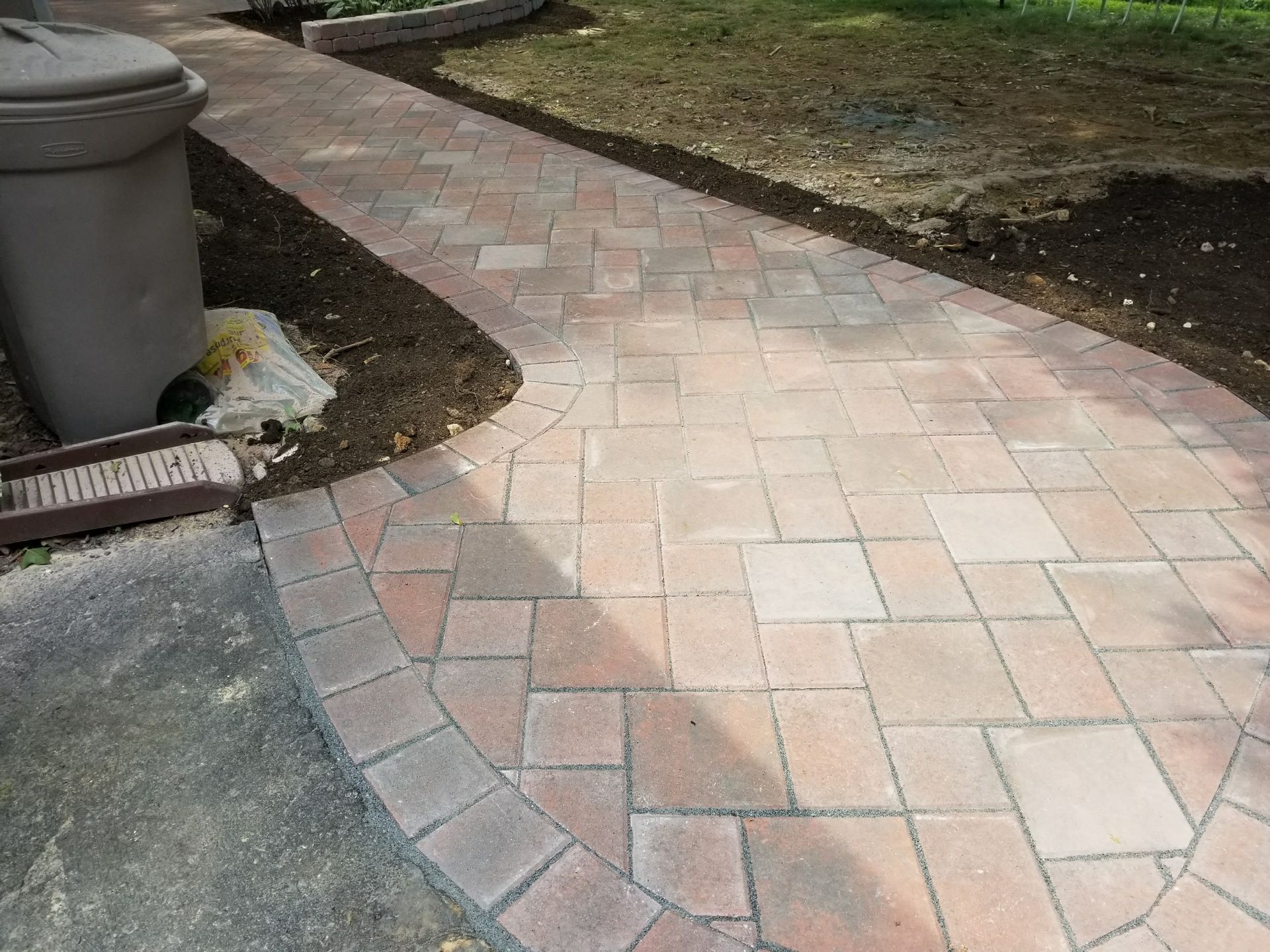 Curved brick walkway with red and tan pavers beside a lawn and trash bin