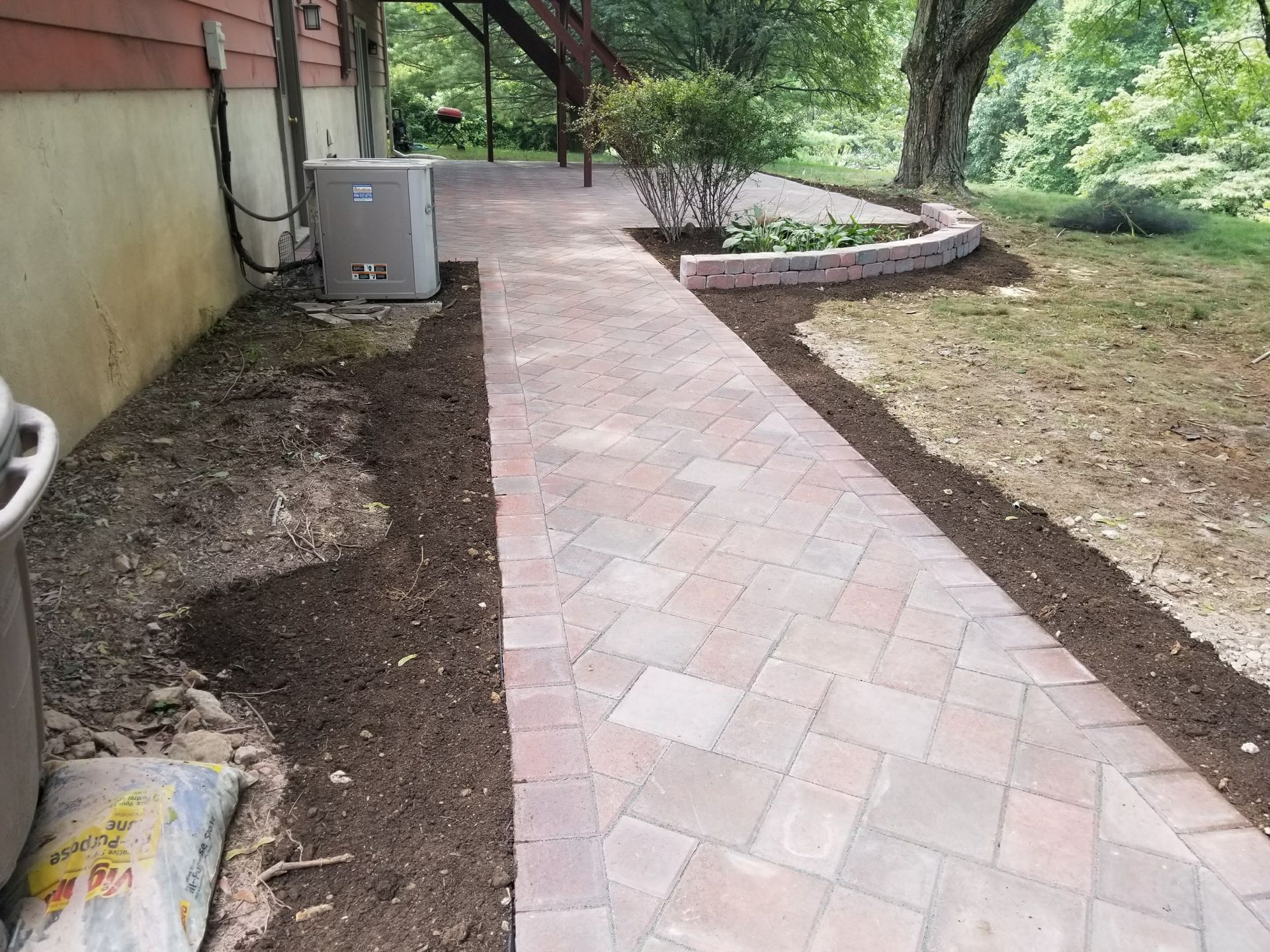 Brick walkway beside a house, bordered by fresh dirt and a grassy yard with trees.