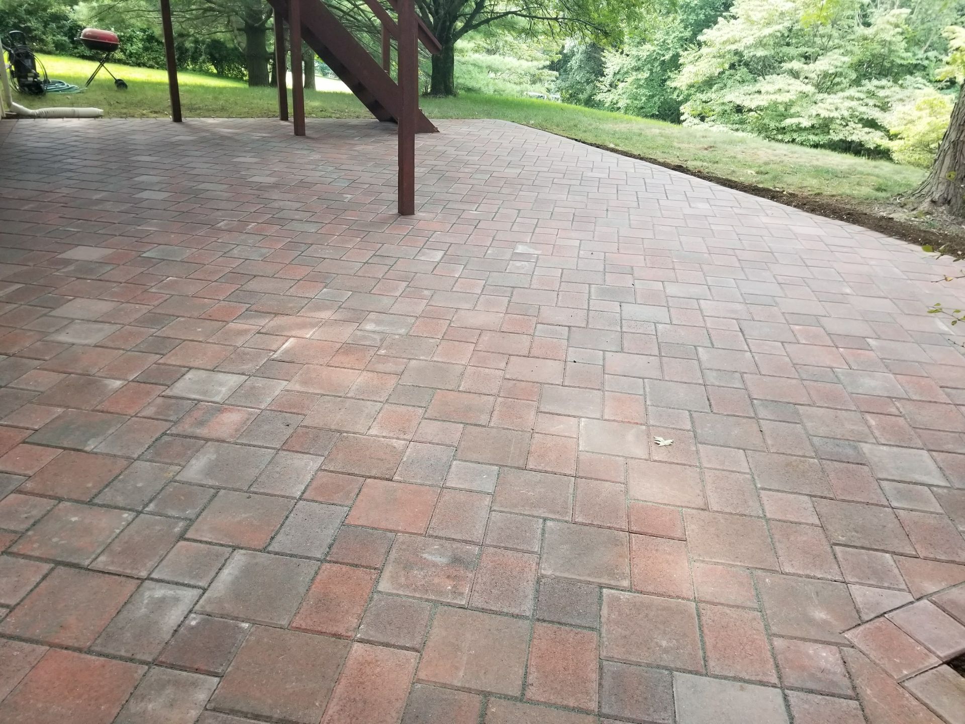 Brick patio beside a grassy park path, with trees and a wooden structure in the background.