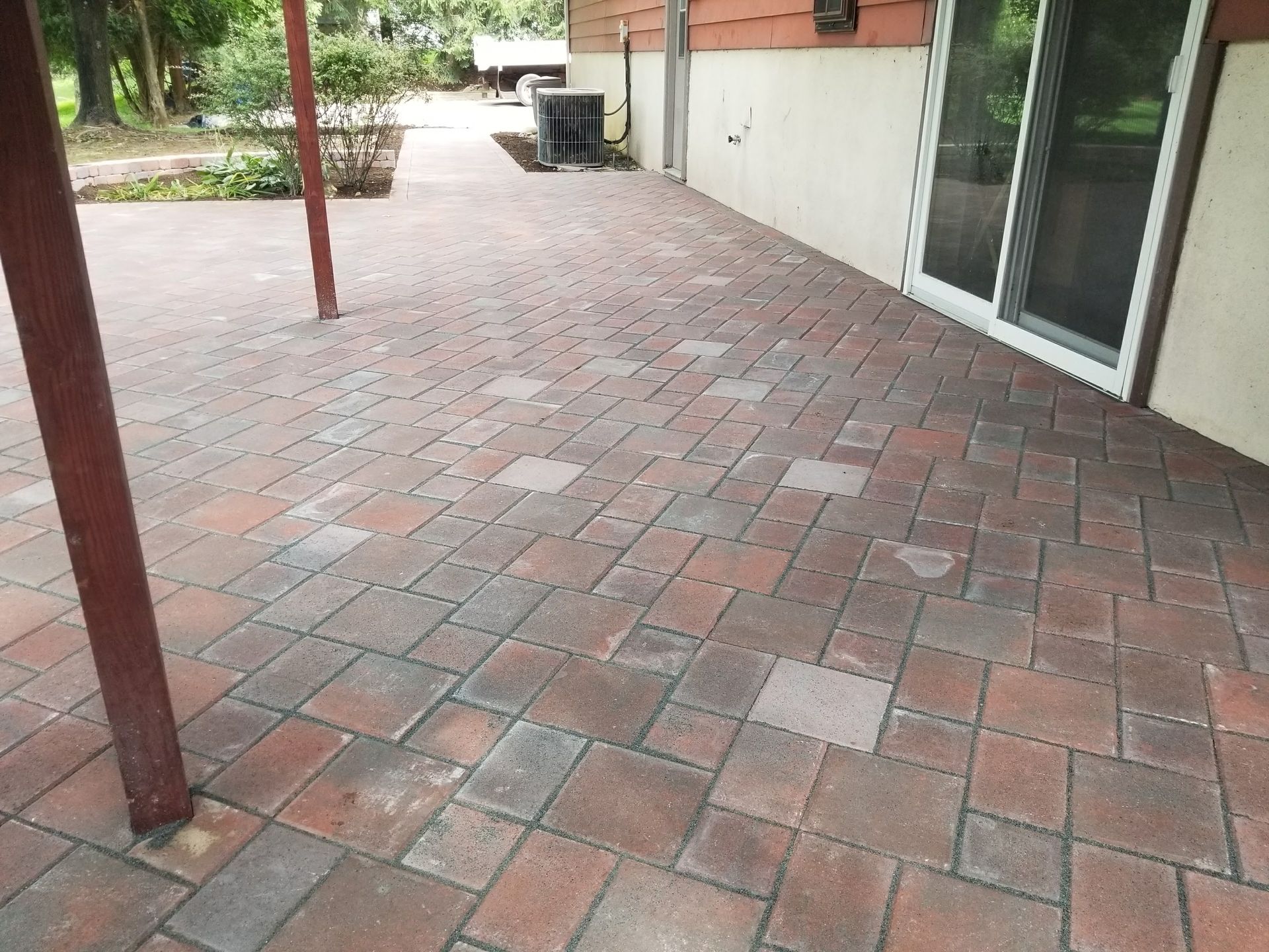 Brick patio beside a house with glass doors and a red support post, leading to a garden area.