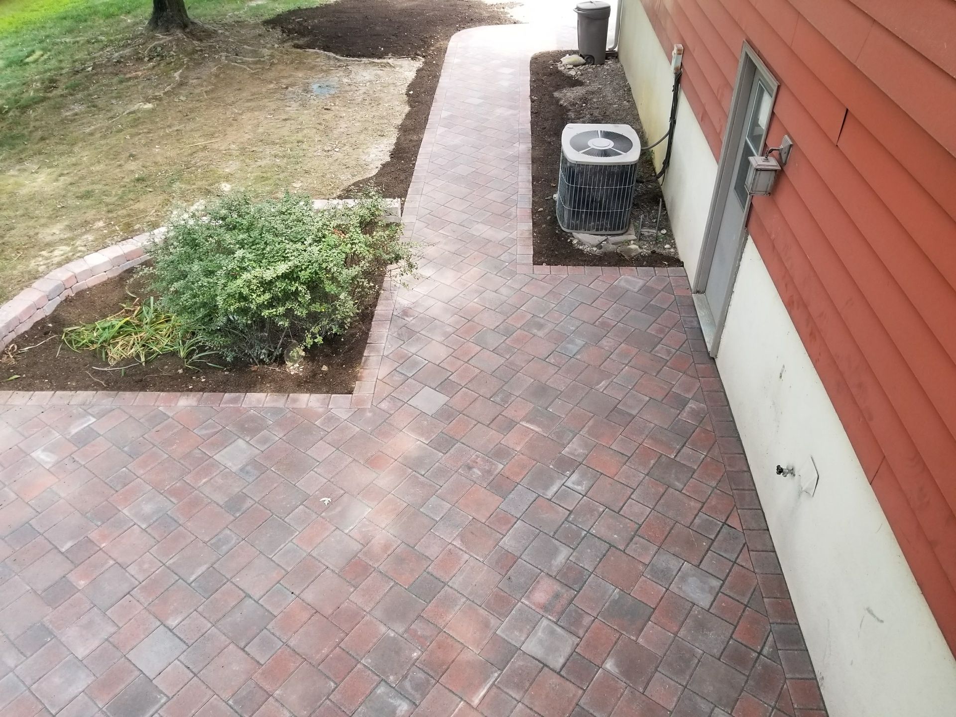 Brick patio walkway beside a red house with an air conditioner and small shrubs.