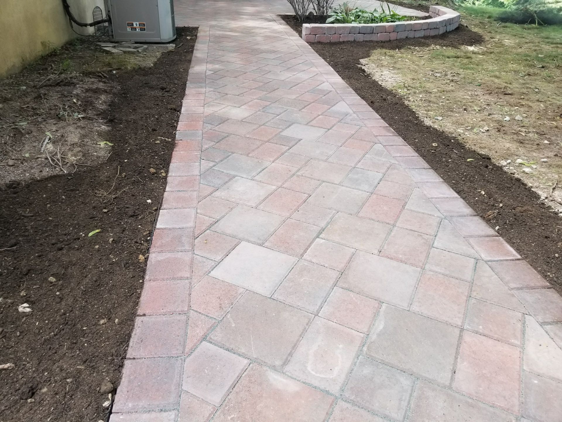 Brick-paved garden walkway bordered by bare soil and grass leading to a patio