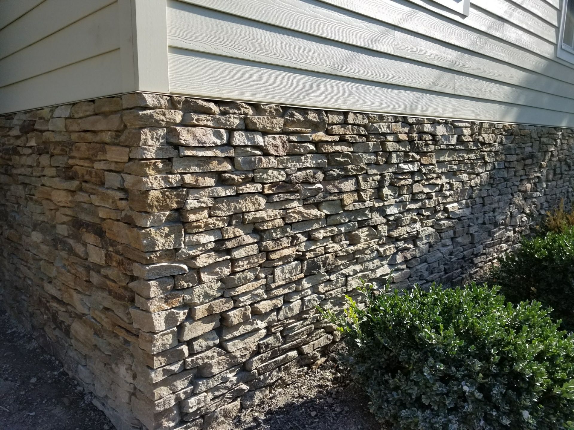 Stone veneer foundation on a house with white siding and green shrubs along the side.