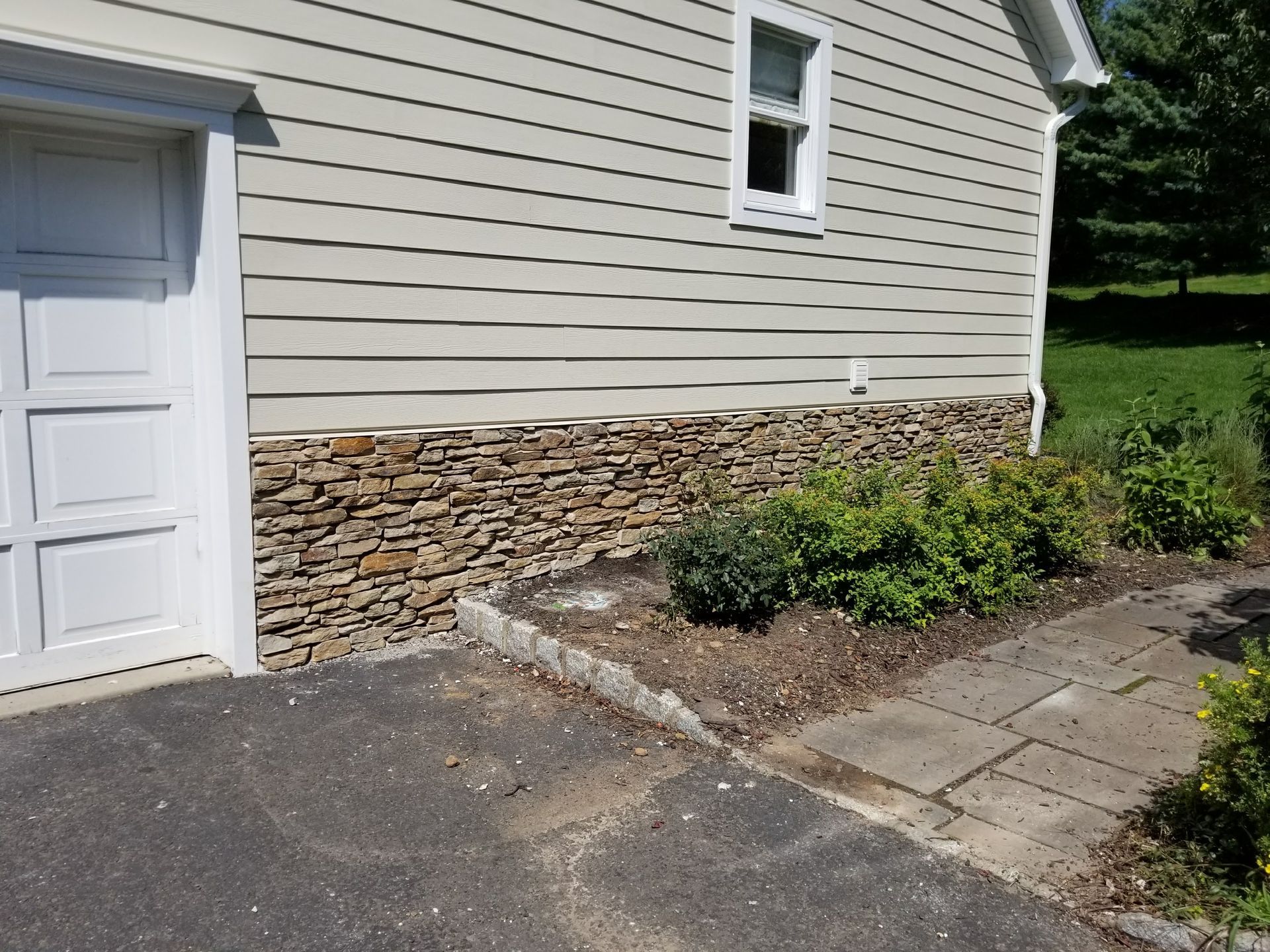 Side of beige house with white door, stone veneer, asphalt driveway, and a small garden bed.