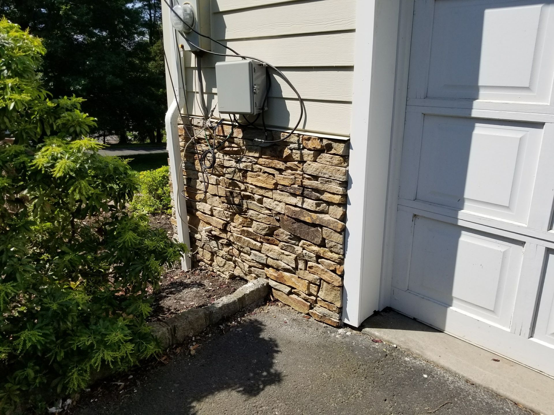 Exterior corner of a house with stone siding, utility box, and a white garage door beside bushes