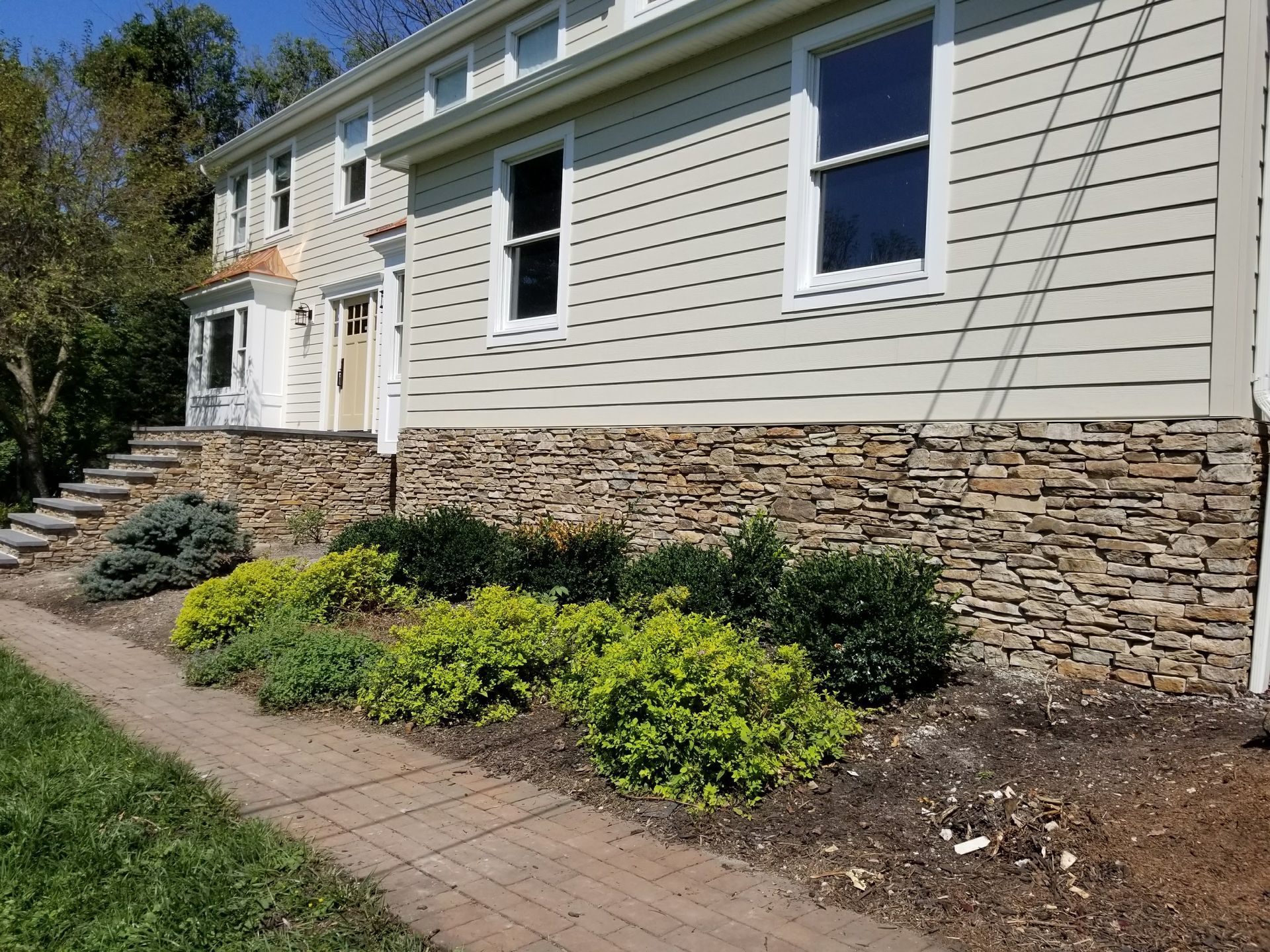 Side view of a beige house with stone foundation, blue window trim, and a landscaped front walkway.