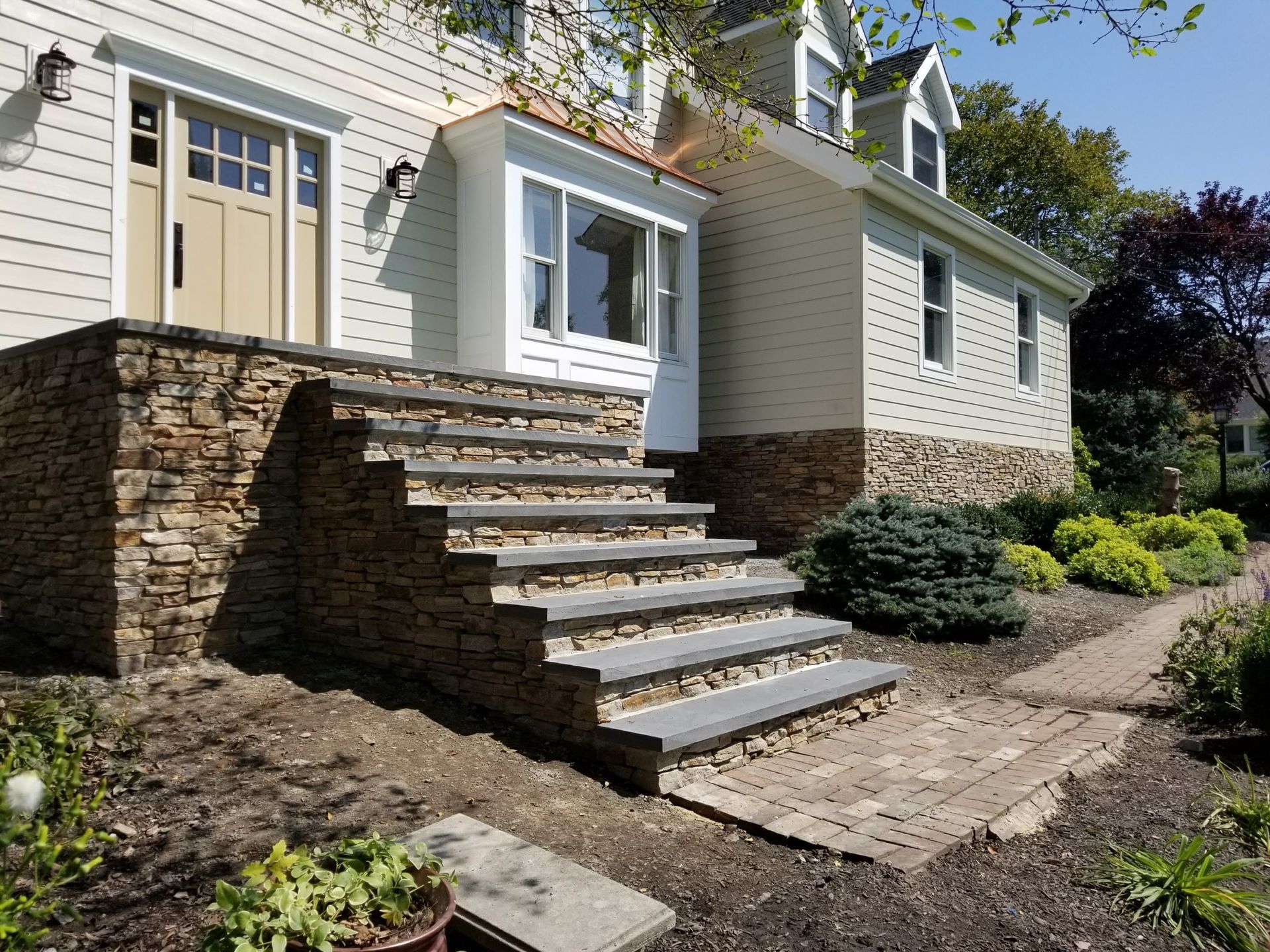 Stone front steps lead to a white house entry with shrubs and a dirt path beside it.