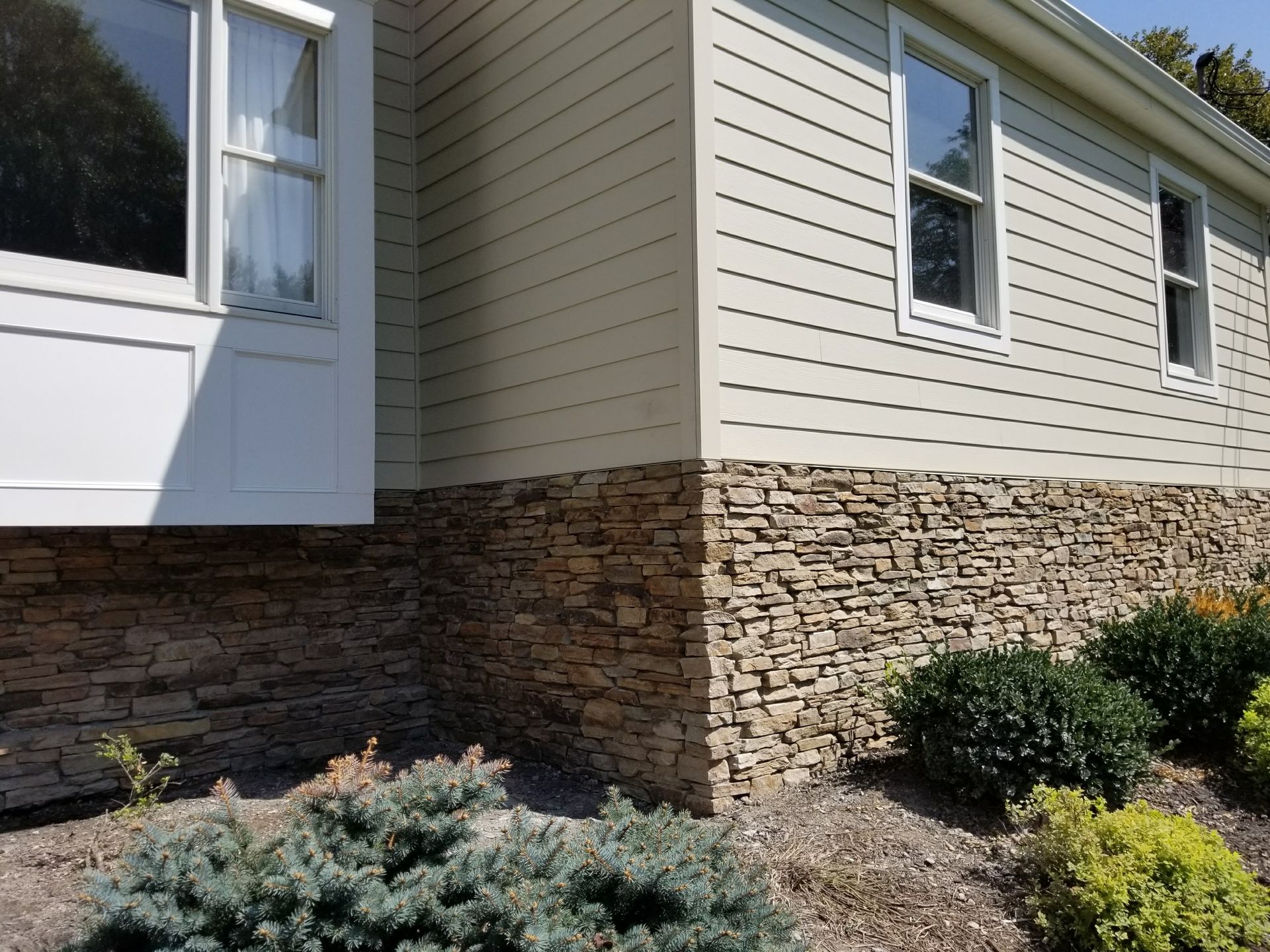 Exterior of a house with beige siding, stone foundation, and landscaped shrubs along the wall
