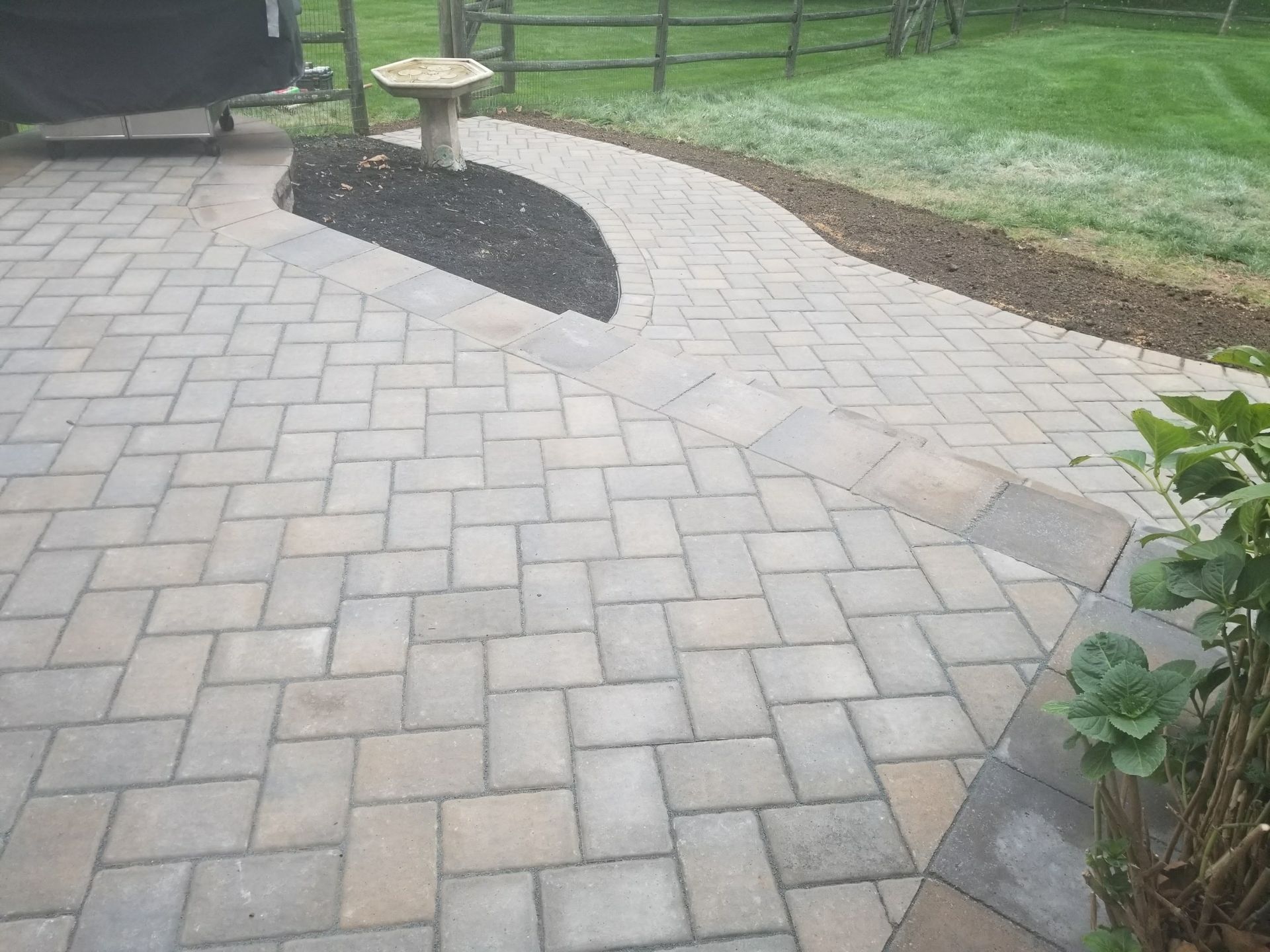 Brick patio path leading to a small stone table and chair in a landscaped yard.