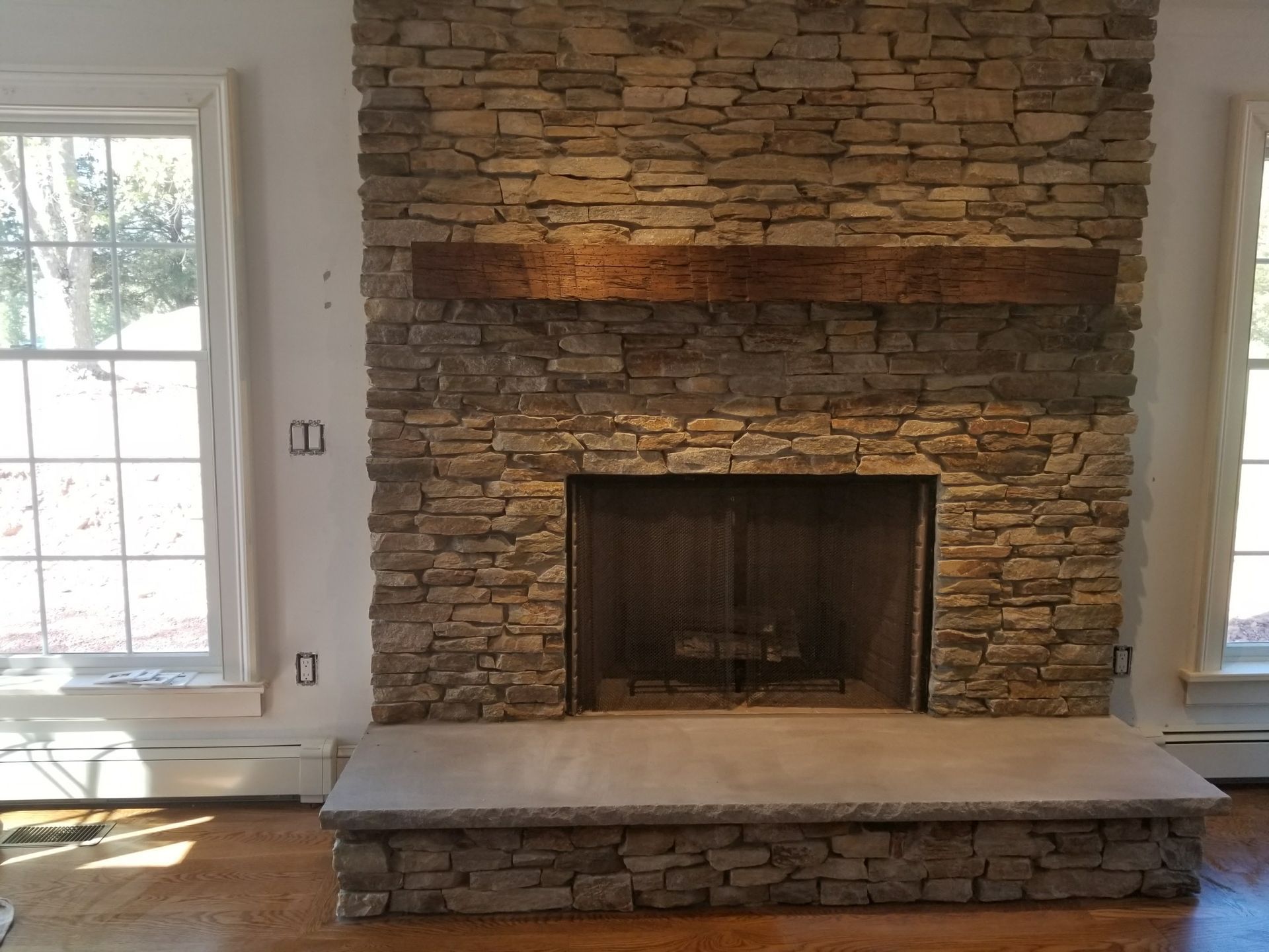 Stone fireplace with a raised hearth in a sunlit room beside two windows.