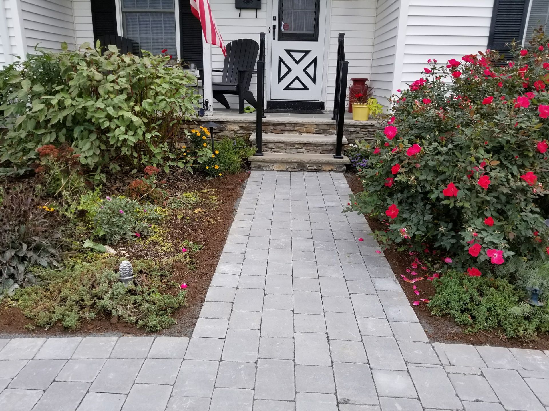 Brick walkway leading to a white porch, bordered by red flowering bushes and green shrubs