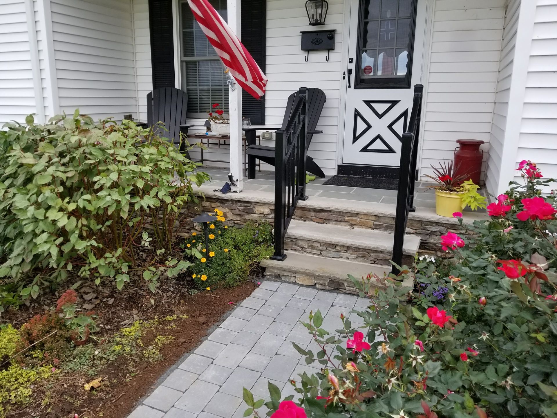 Front porch with black handrails, American flag, and blooming red and yellow flowers by a stone walkway.