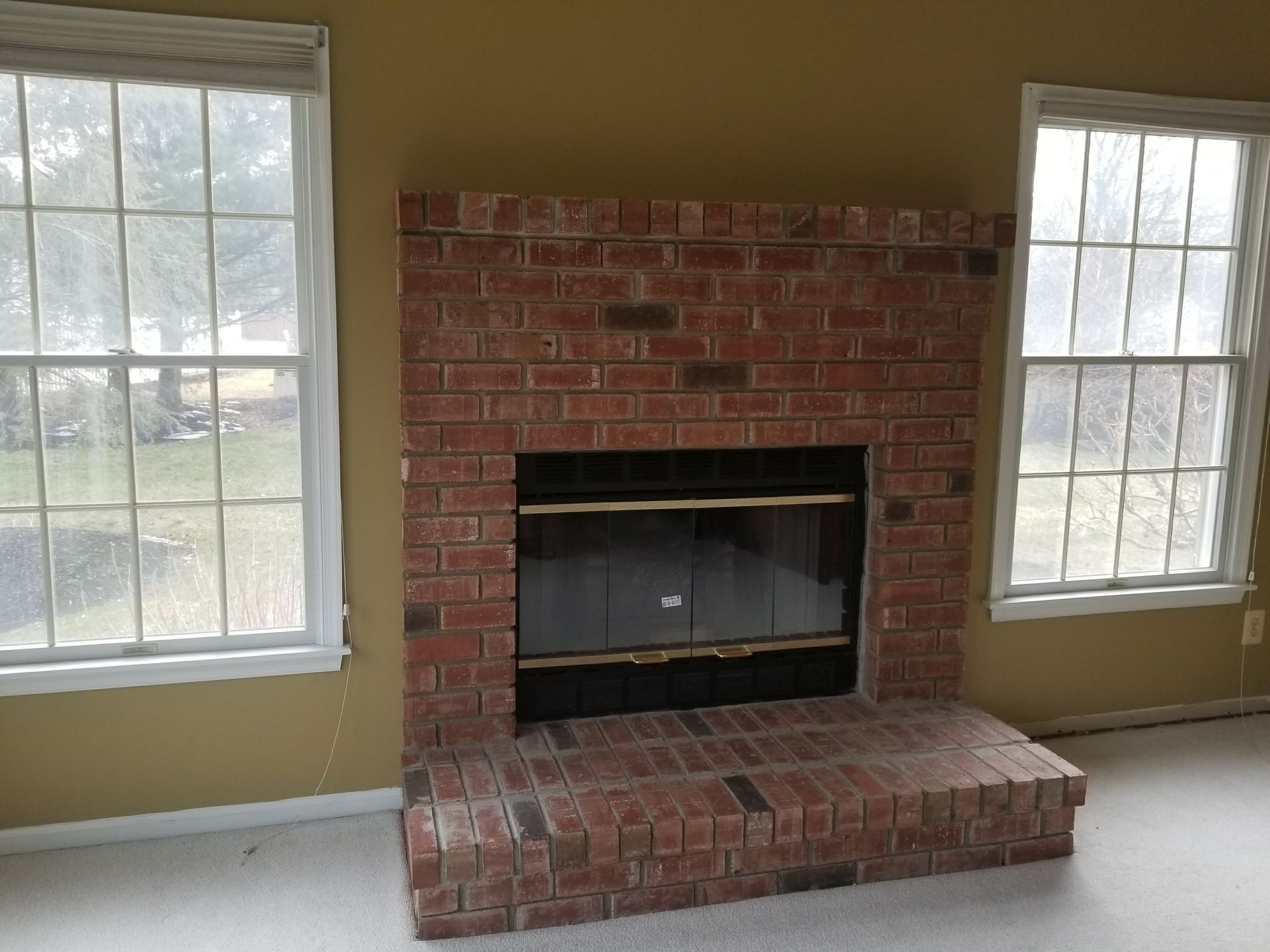 Brick fireplace between two windows in a sparsely furnished room with tan walls and white trim