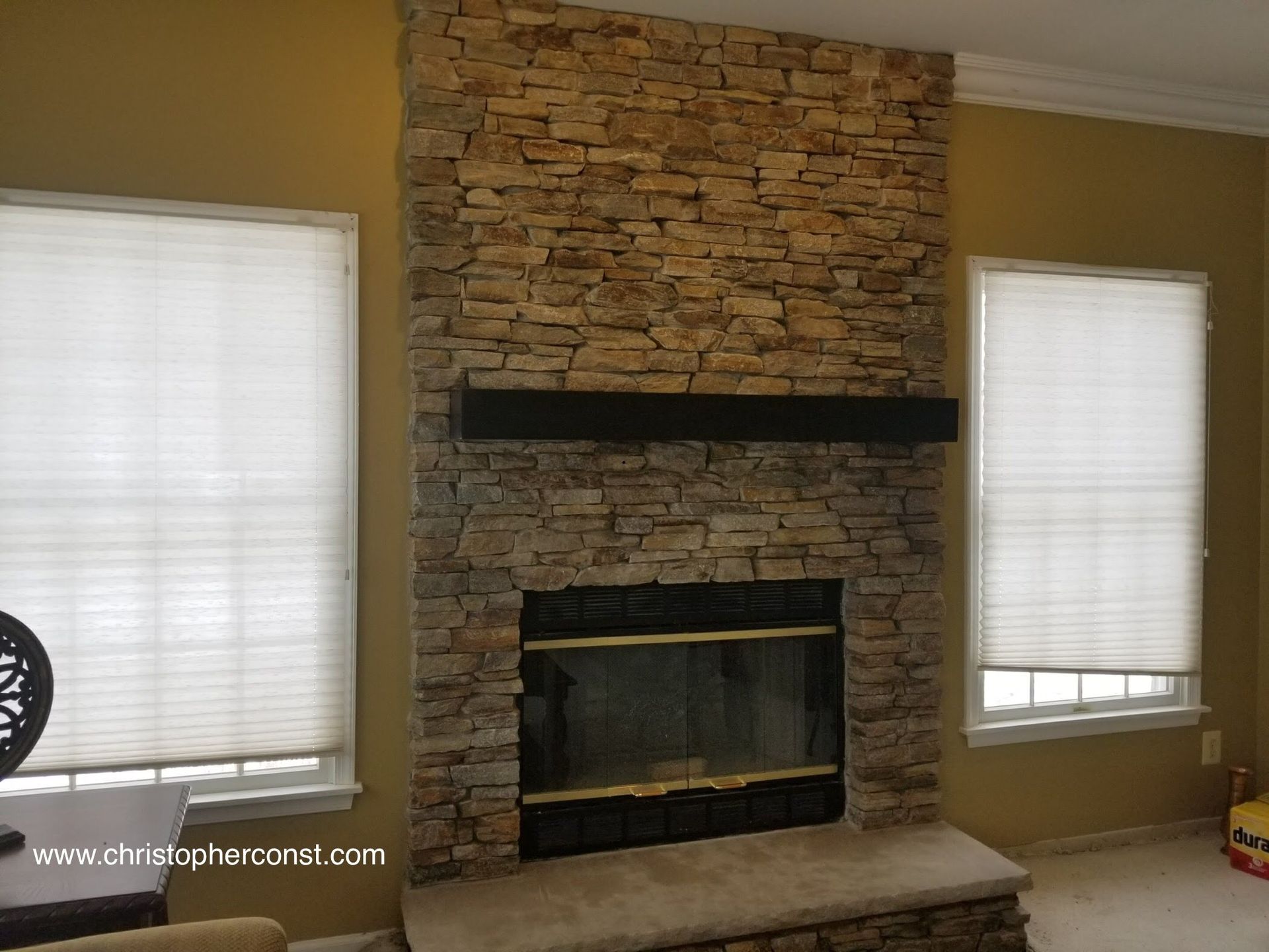 Living room fireplace with stacked stone surround, black mantel, and windows on both sides