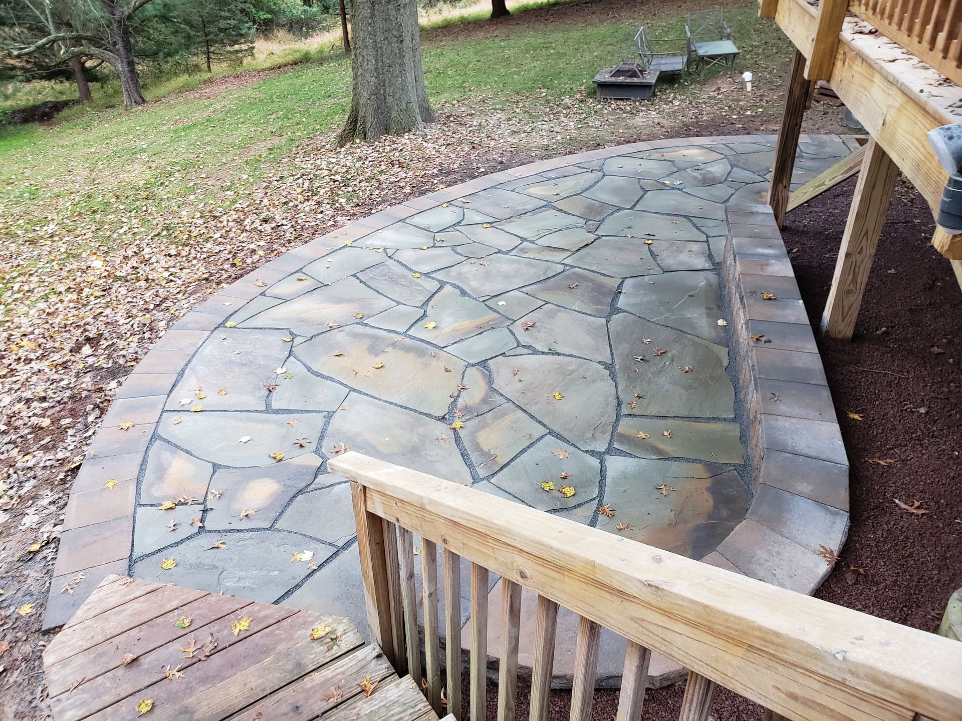 Curved stone patio with wooden deck railing and grassy yard under trees