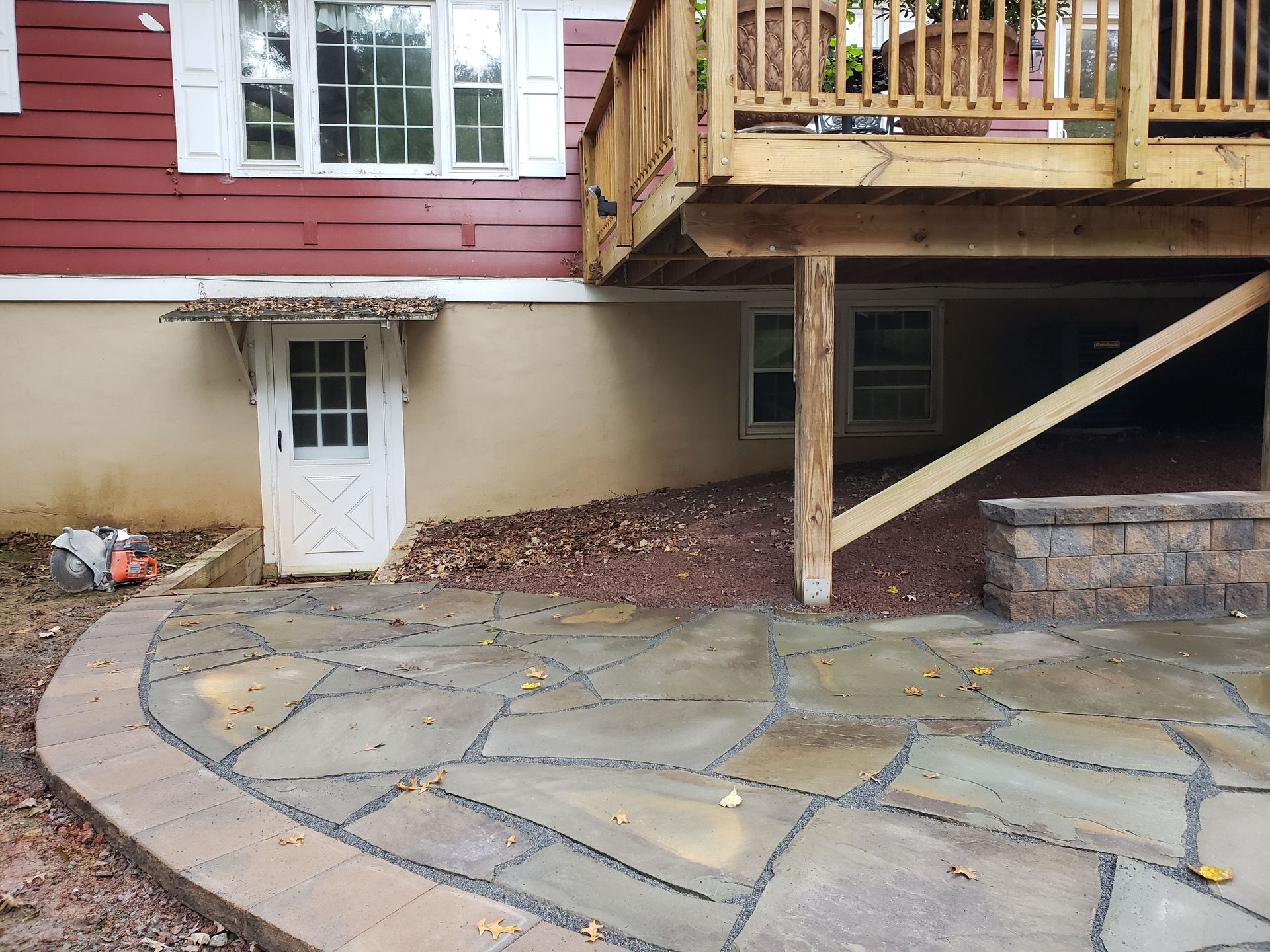 Stone patio beside red house with basement door, brick steps, and raised wooden deck above.