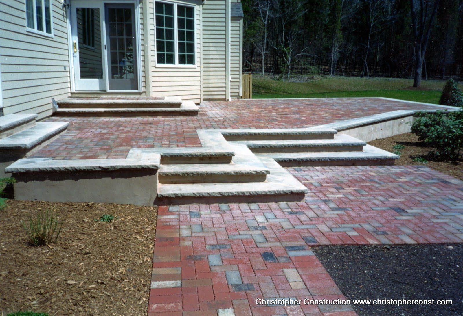 Brick patio and steps leading to a house entrance, with a raised wooden deck and landscaped edges.