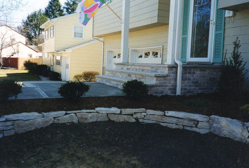 Front yard with stone border, small shrubs, and beige suburban house with a flag near the porch.