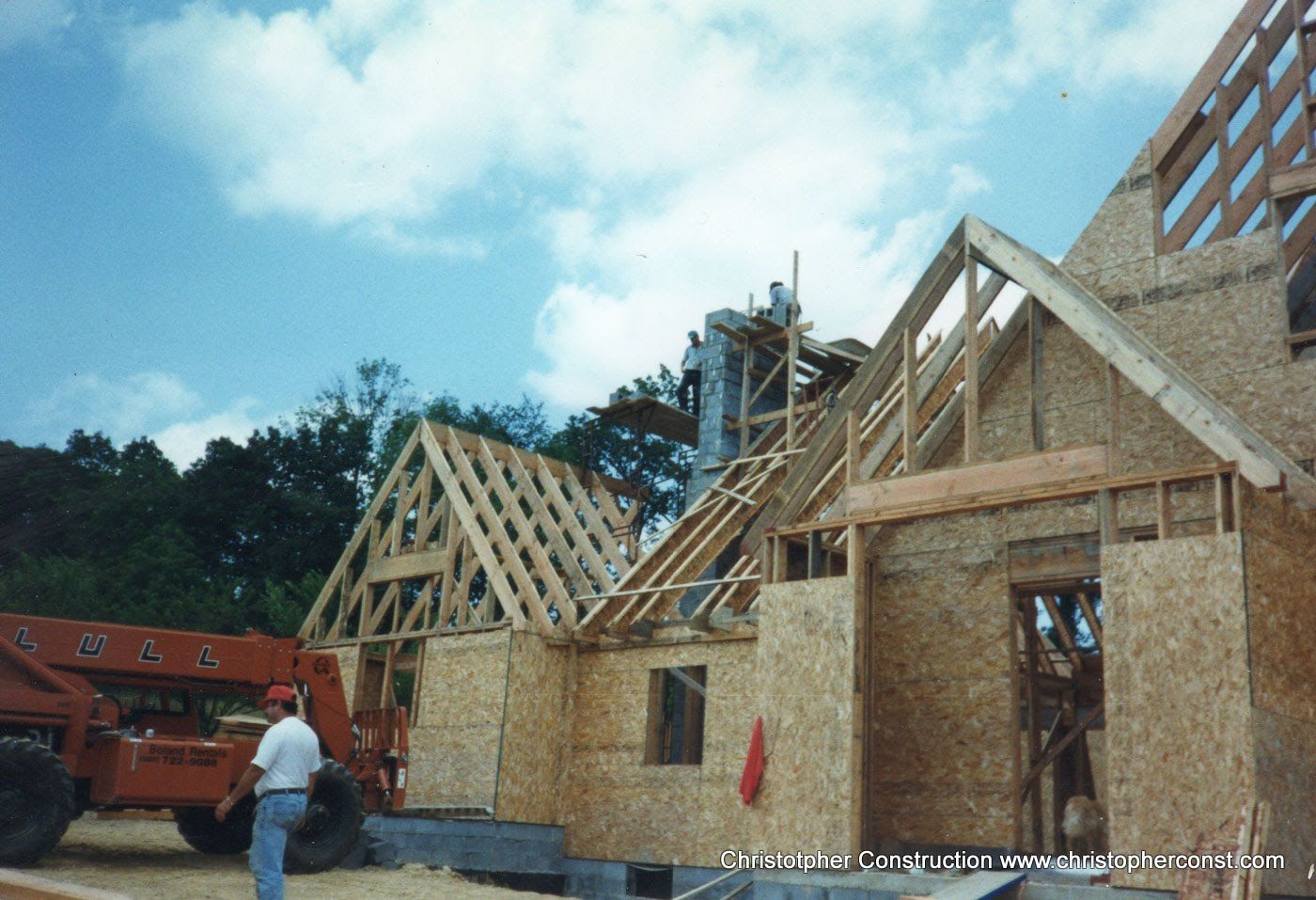 House under construction with exposed roof trusses, a worker, and a truck on site under a blue sky